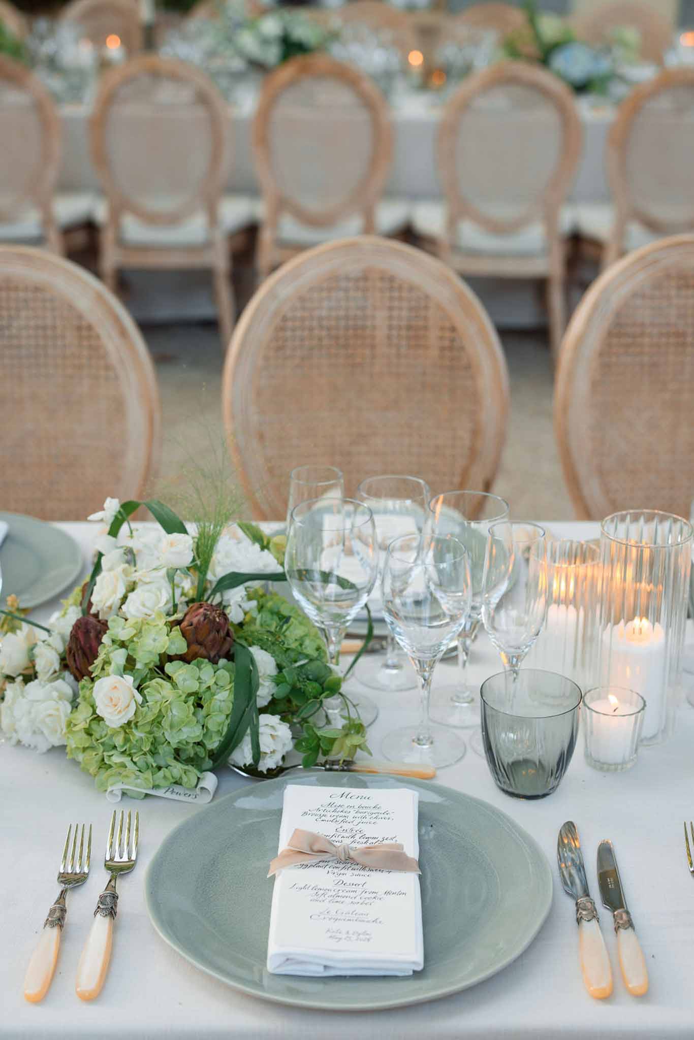 Reception table setting with sage charger plate, floral centerpiece, and candles at wedding venue