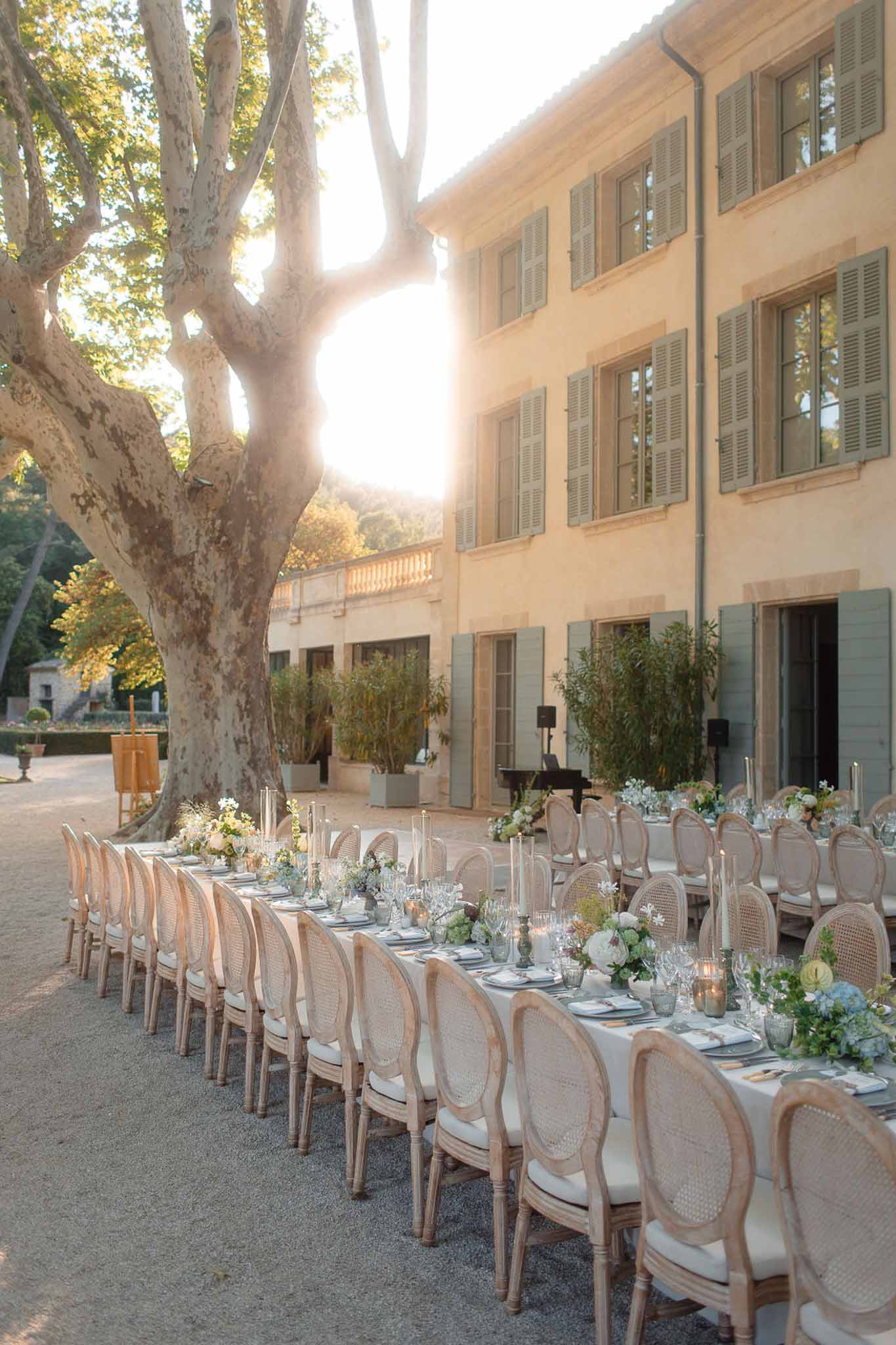 Long reception table with ivory linens and floral centerpieces in villa courtyard beneath mature tree