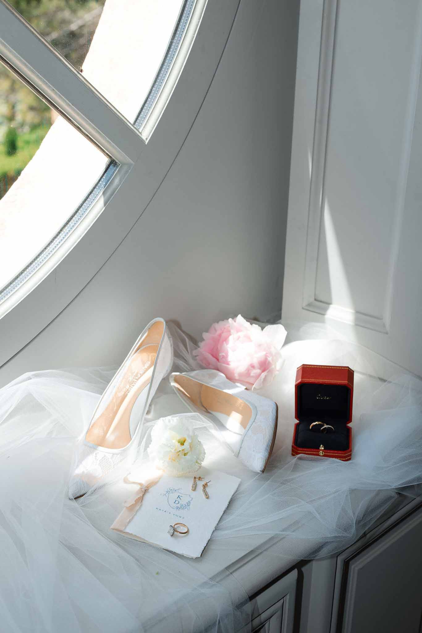 Bridal flat lay with shoes, veil, rings and pink peonies arranged on blue cushion by window