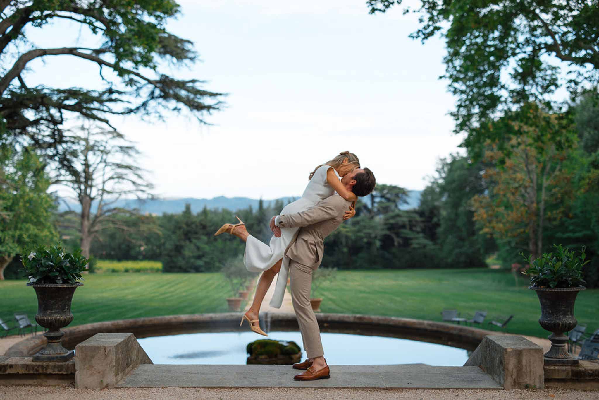 Groom lifting bride on stone bridge over reflecting pool at estate garden