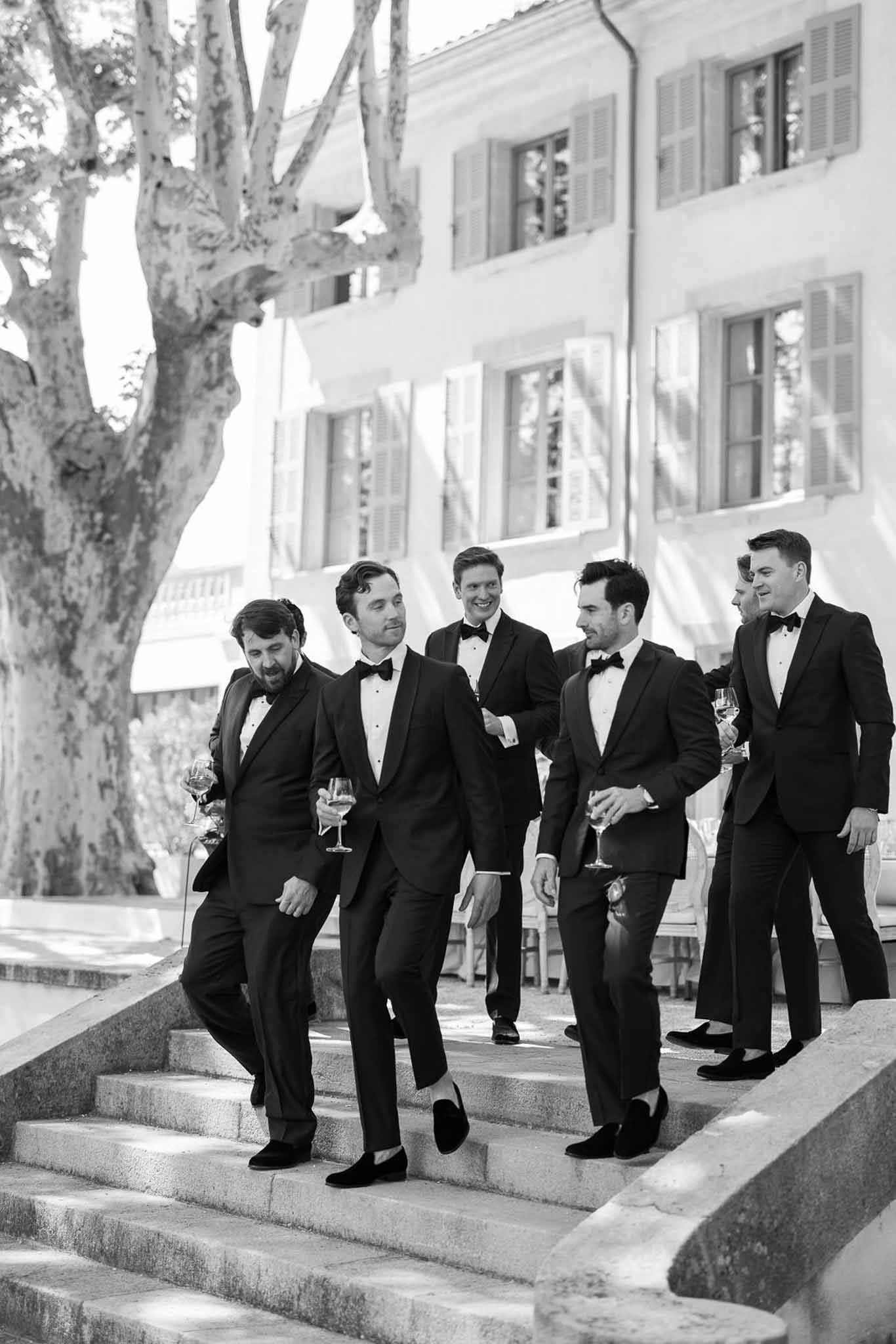 Groom and groomsmen in black tuxedos walking with champagne at outdoor wedding venue