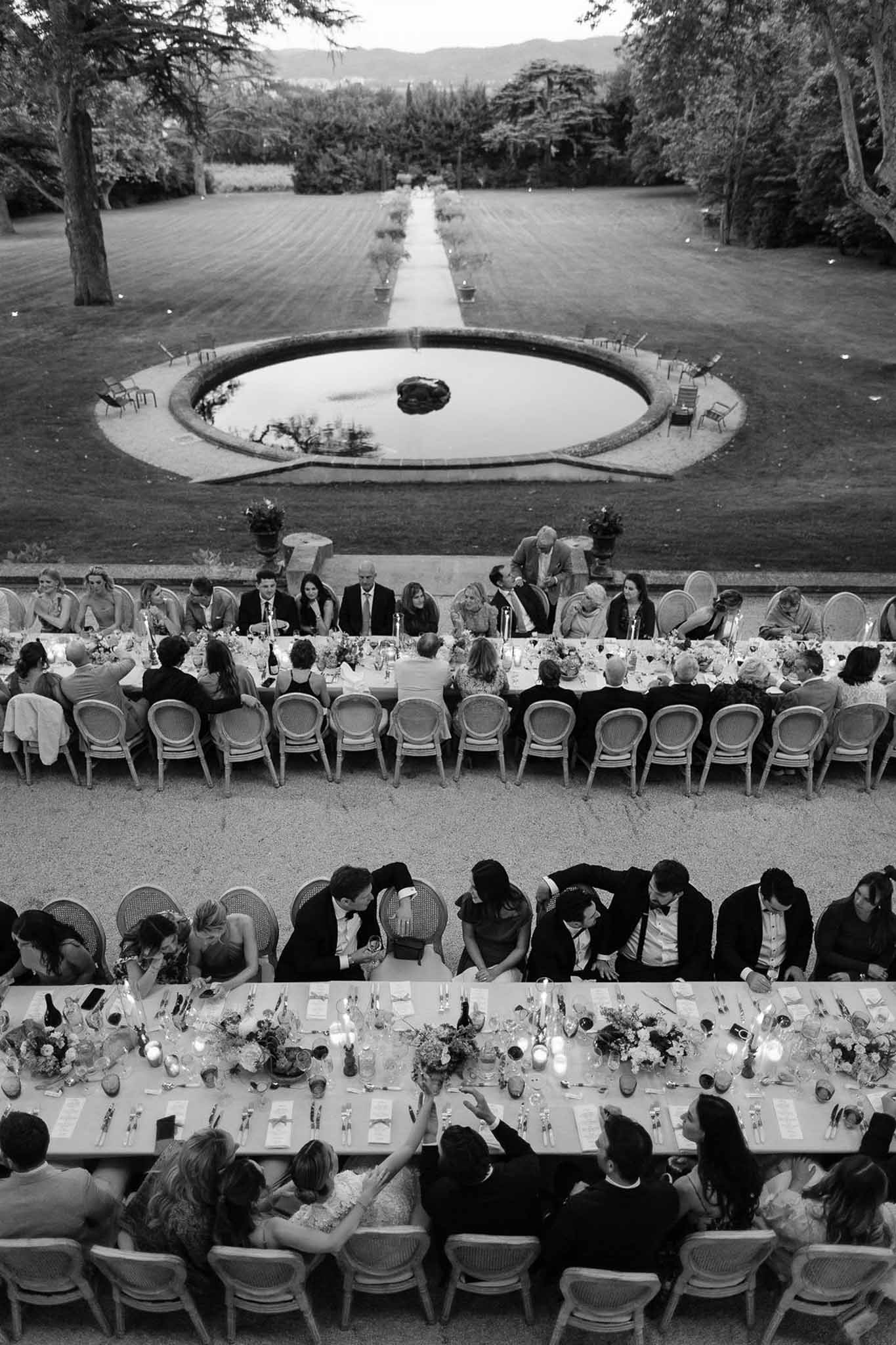 Aerial view of outdoor wedding reception with long tables and water feature in garden setting