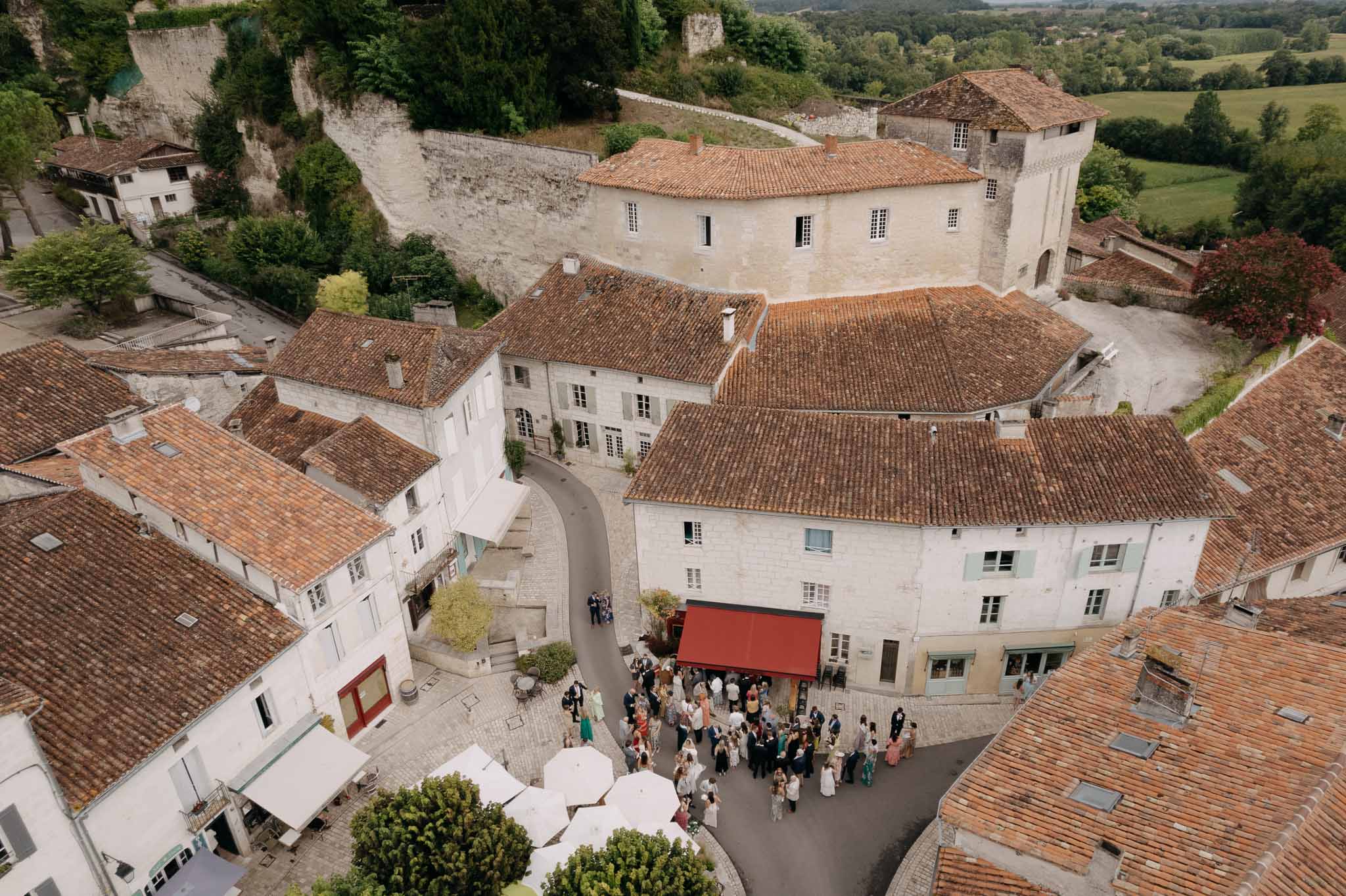 Aerial view of wedding cocktail hour in a historic French village courtyard with 60-80 guests mingling among limestone bui...