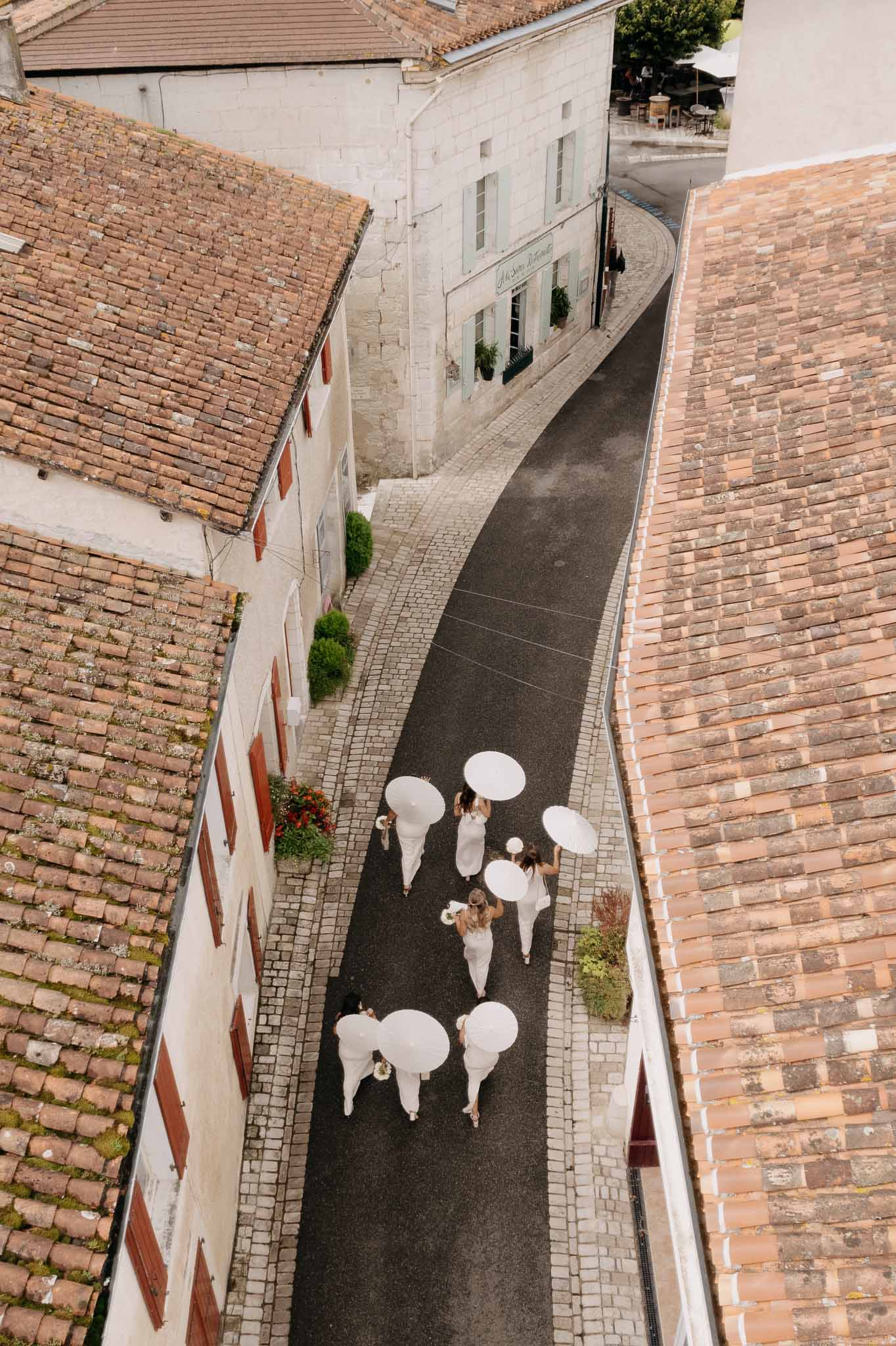 Aerial view of bridal party in white gowns carrying white parasols on winding village street