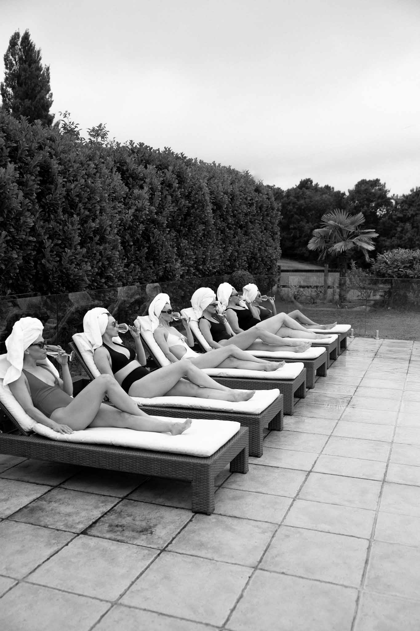 Black and white photo of bridesmaids in towel wraps sipping champagne on poolside sun loungers