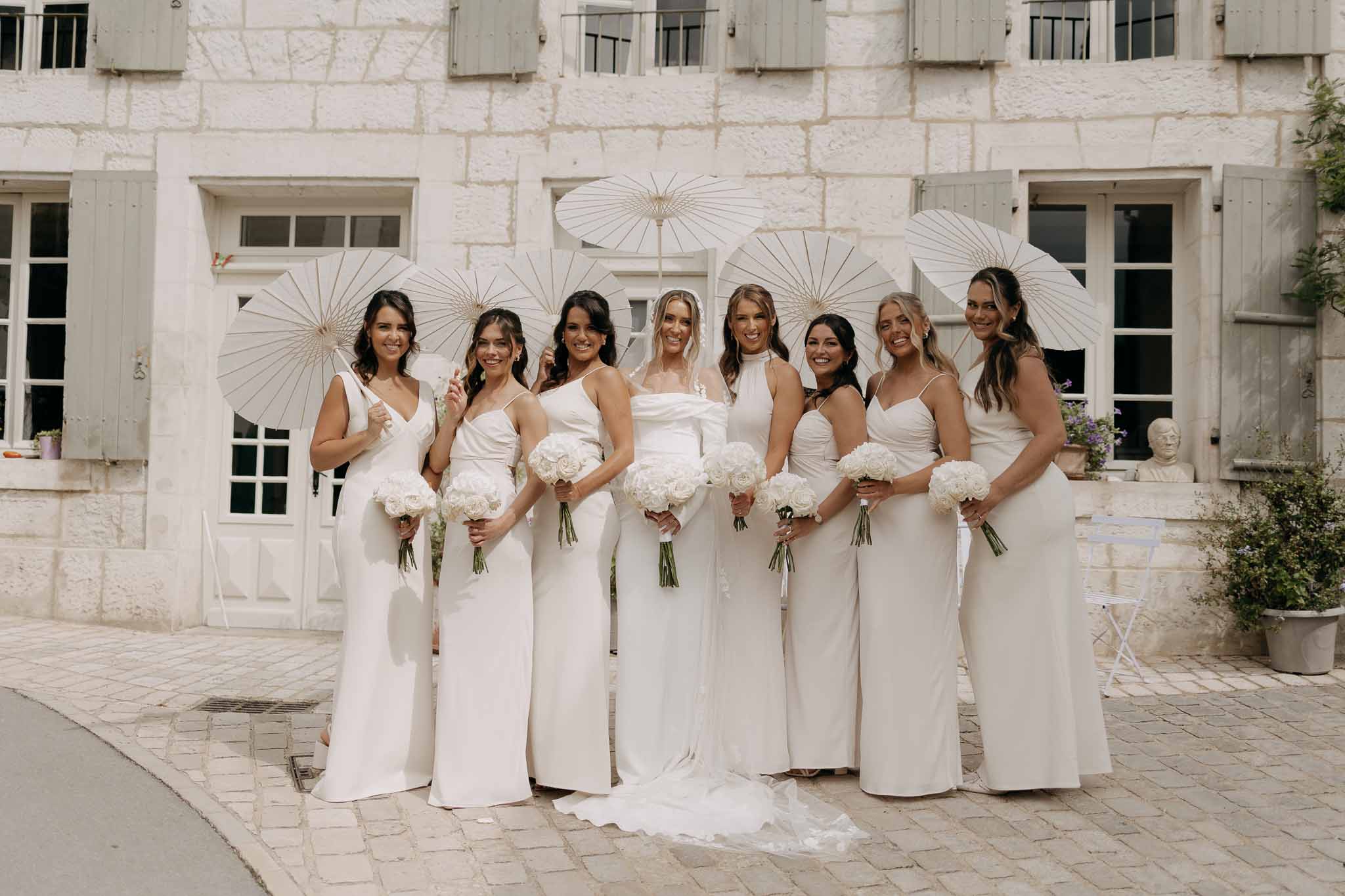Bride and seven bridesmaids in white and ivory gowns holding bouquets and parasols outside a limestone manor