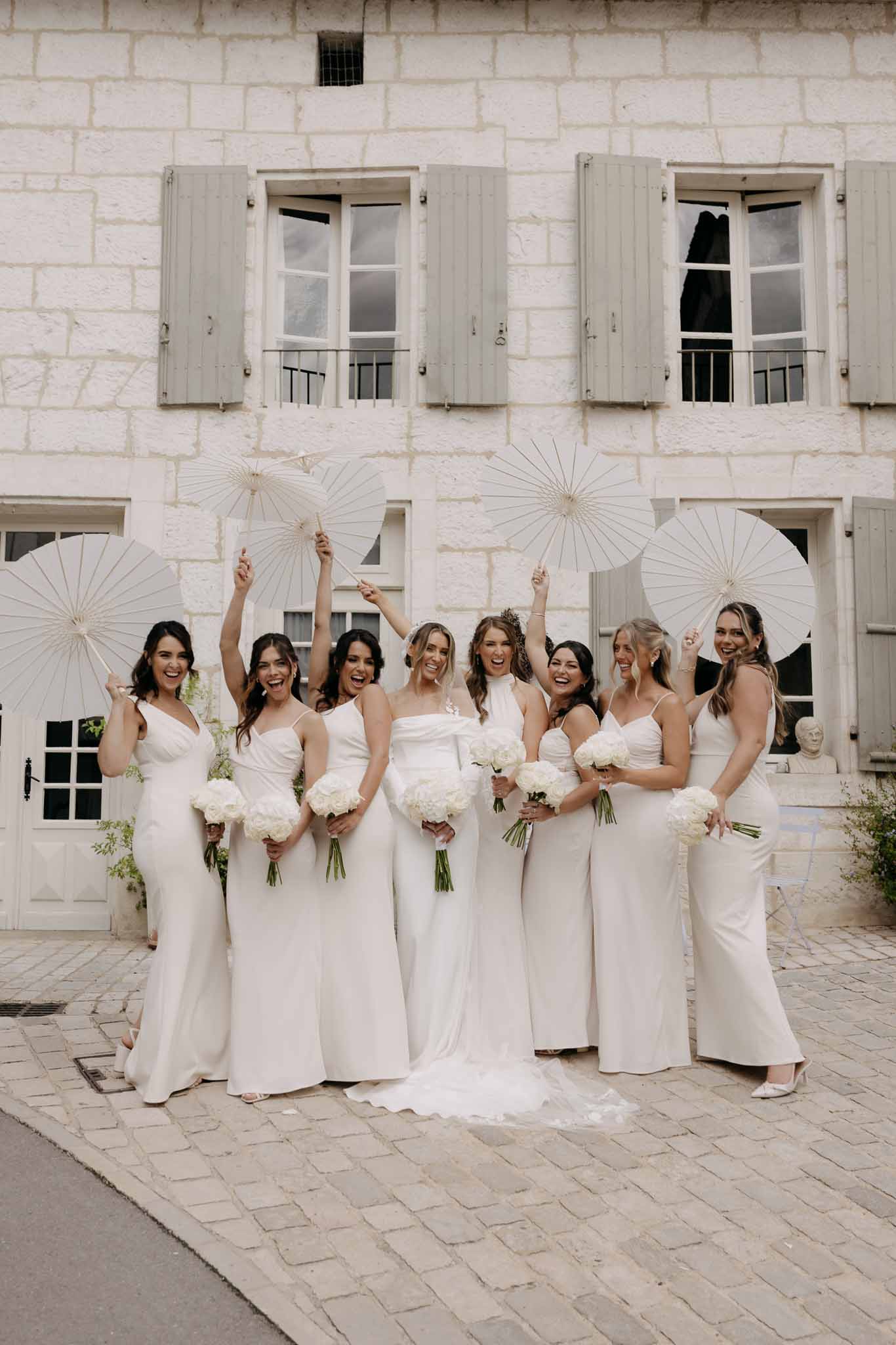 Bride and six bridesmaids in ivory slip dresses raising white parasols with peony bouquets
