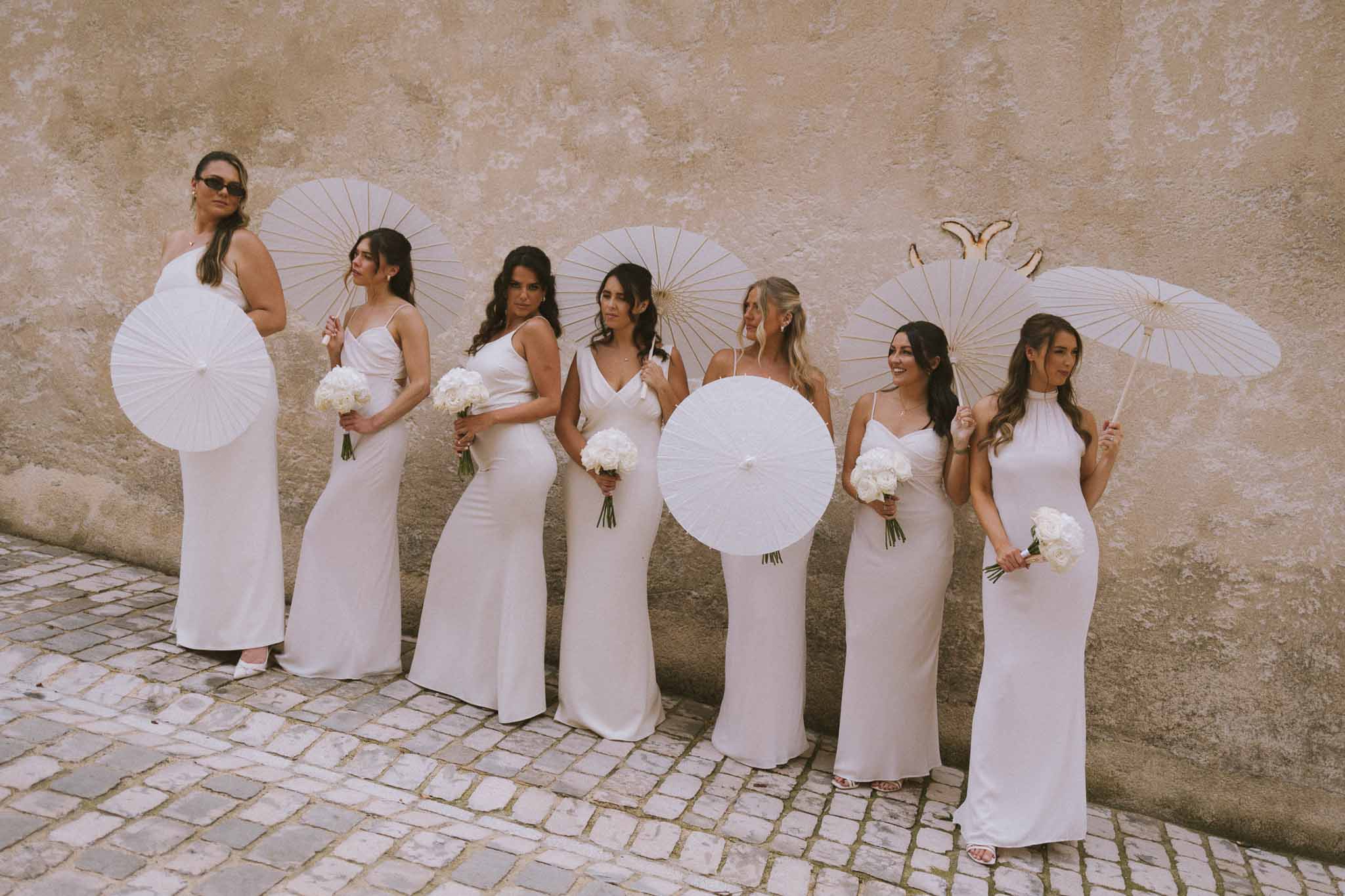 Seven women in white slip dresses holding parasols and peony bouquets against ochre stone wall