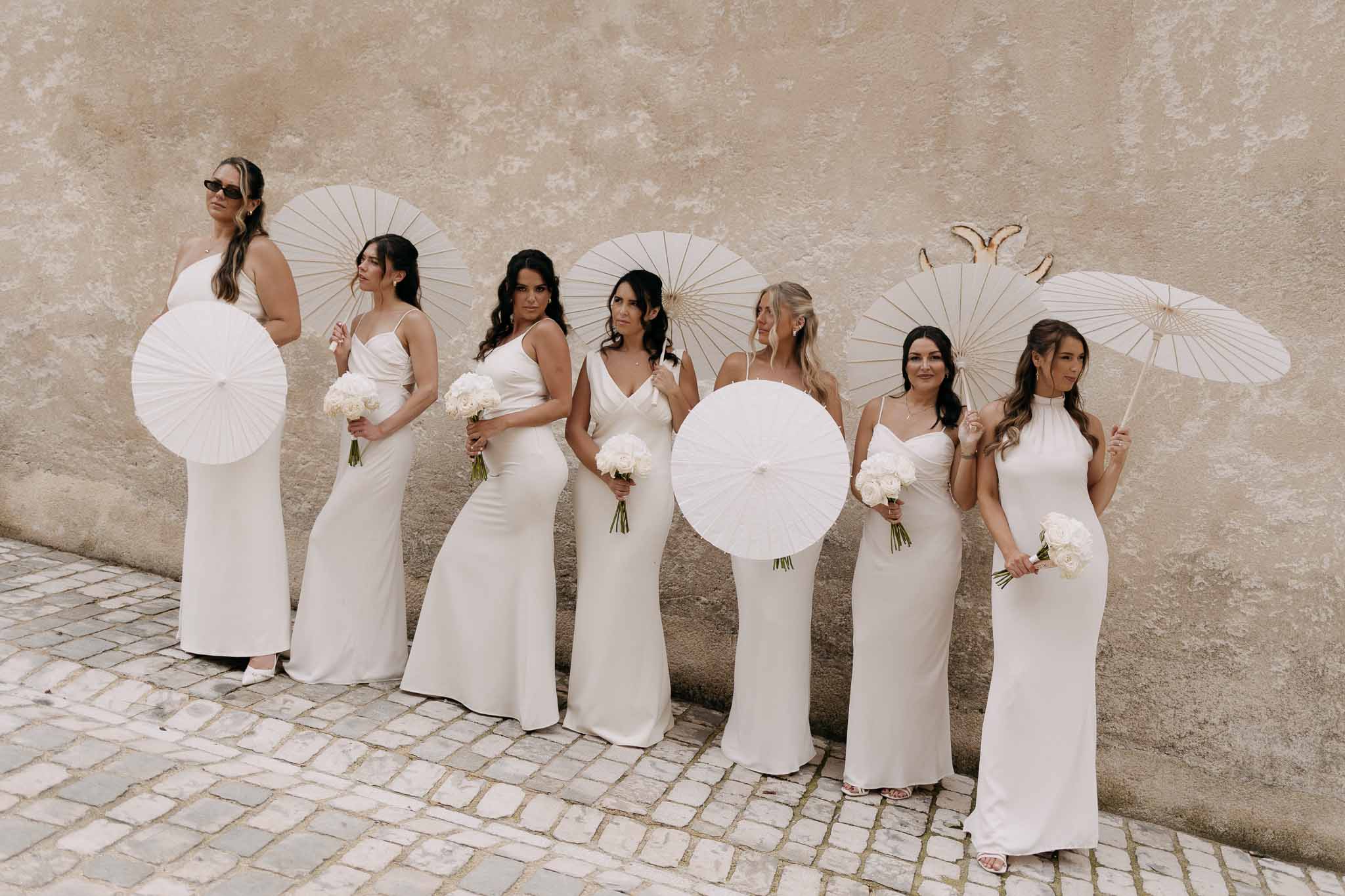 Seven women in ivory mismatched slip gowns with white parasols and peony bouquets against weathered plaster wall