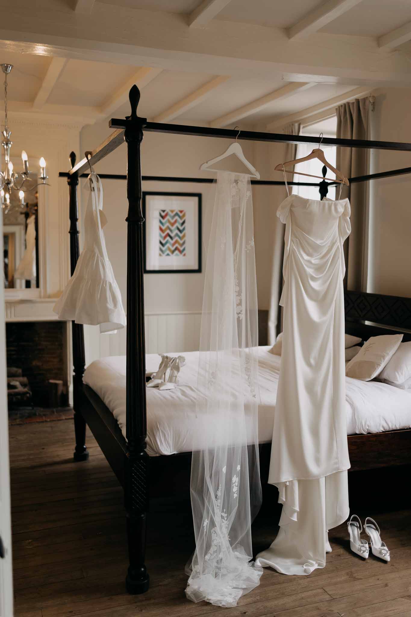 Wedding dress, cathedral-length veil, and mini dress hung on black four-poster bed with bridal heels on wooden floor