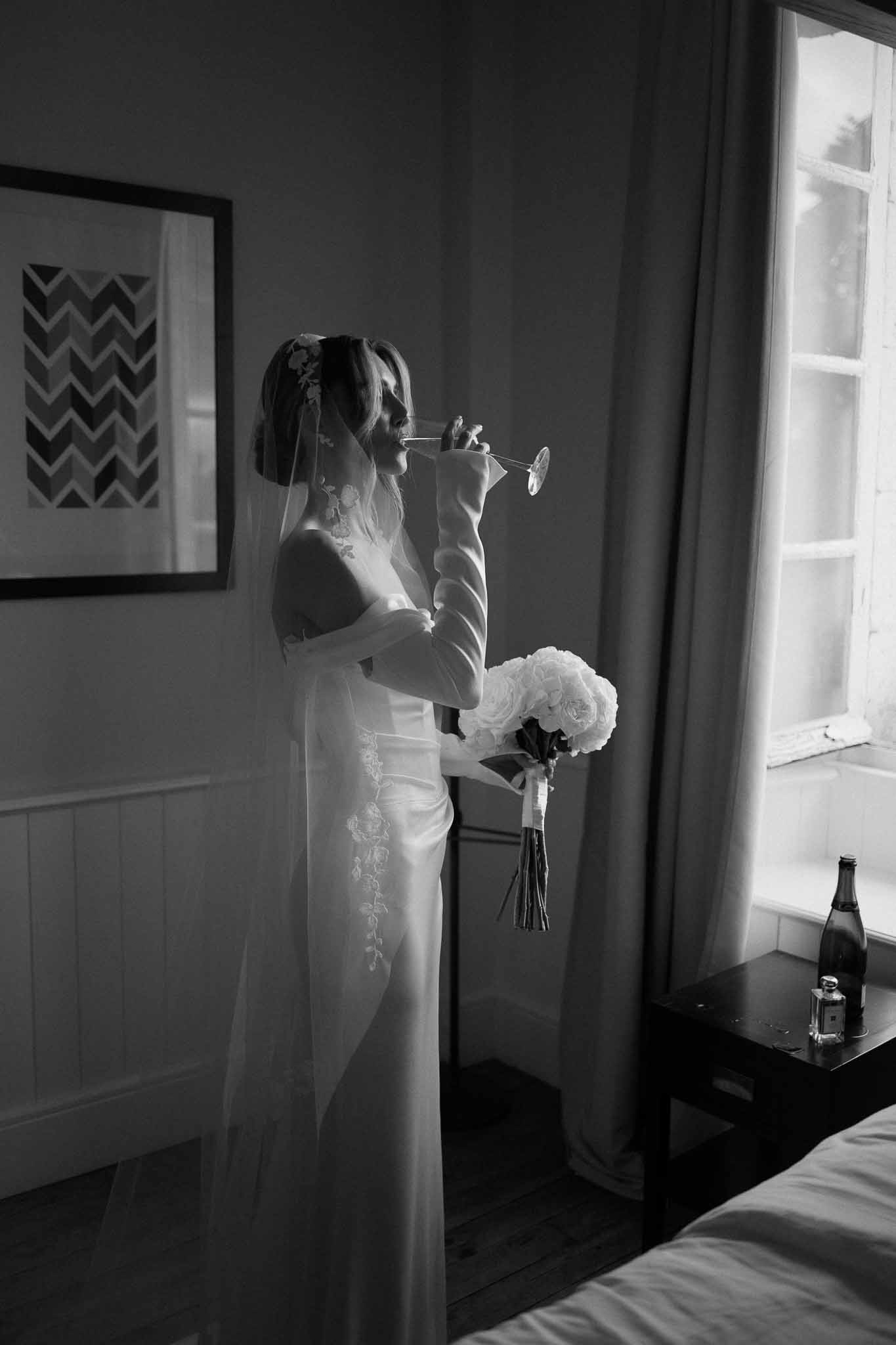 Black and white portrait of bride sipping champagne by a window wearing lace gown, opera gloves, and cathedral veil