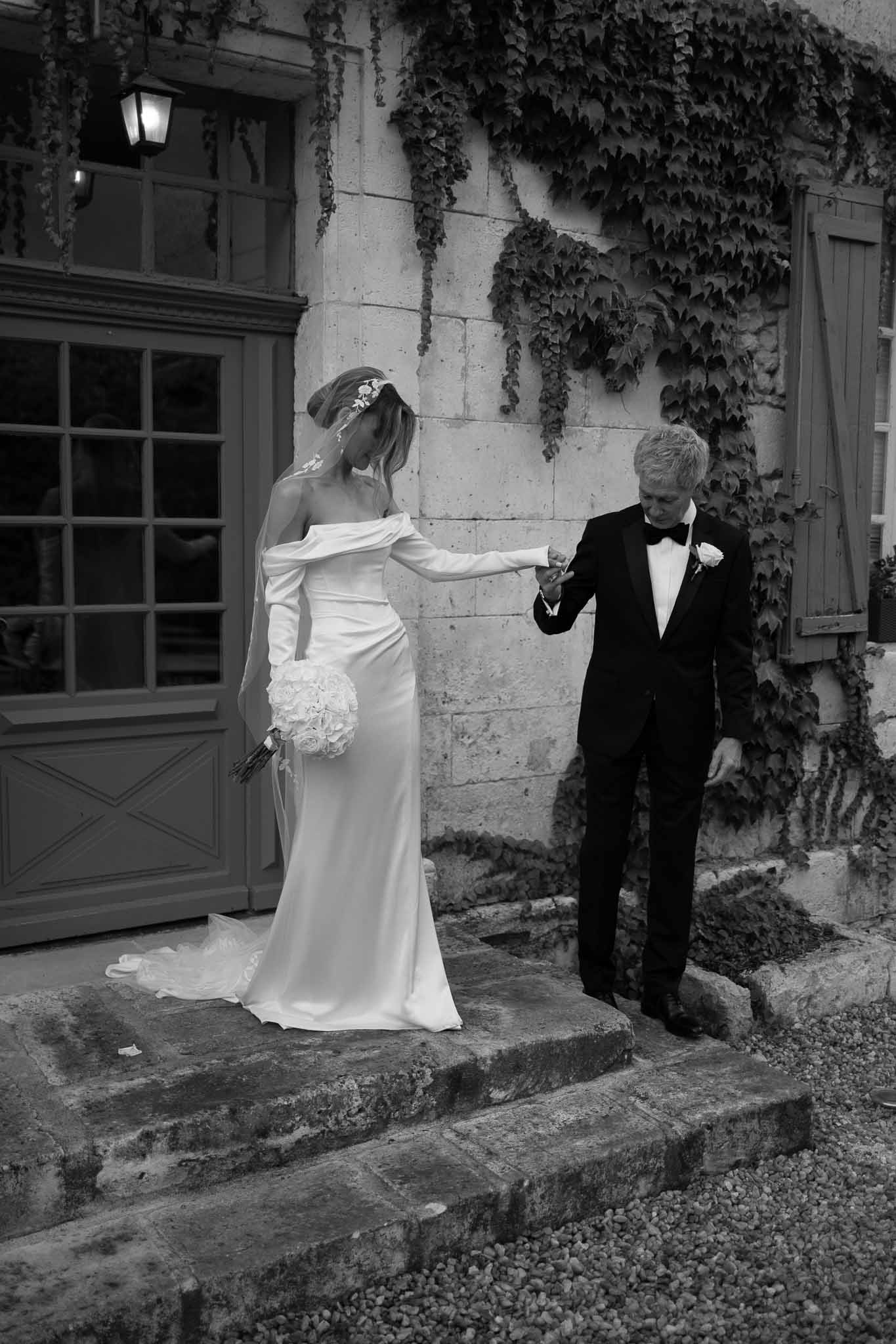 Black and white groom holding bride's hand descending ivy-covered chateau steps with rose bouquet and lace train