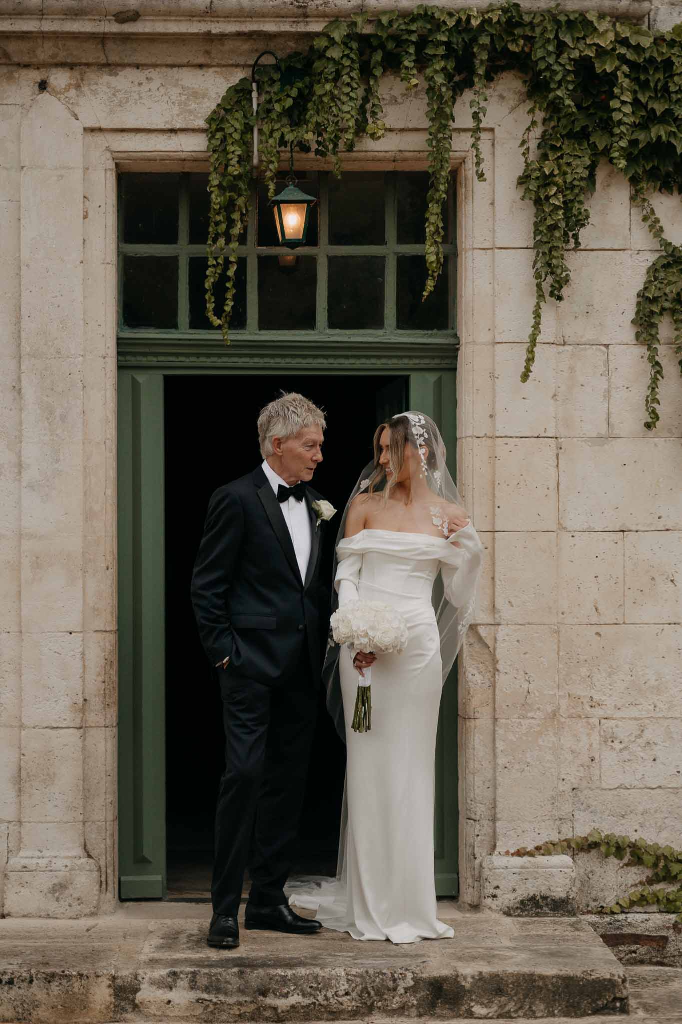 Bride with cathedral veil and white peony bouquet beside father in tuxedo in green-doored chateau entrance