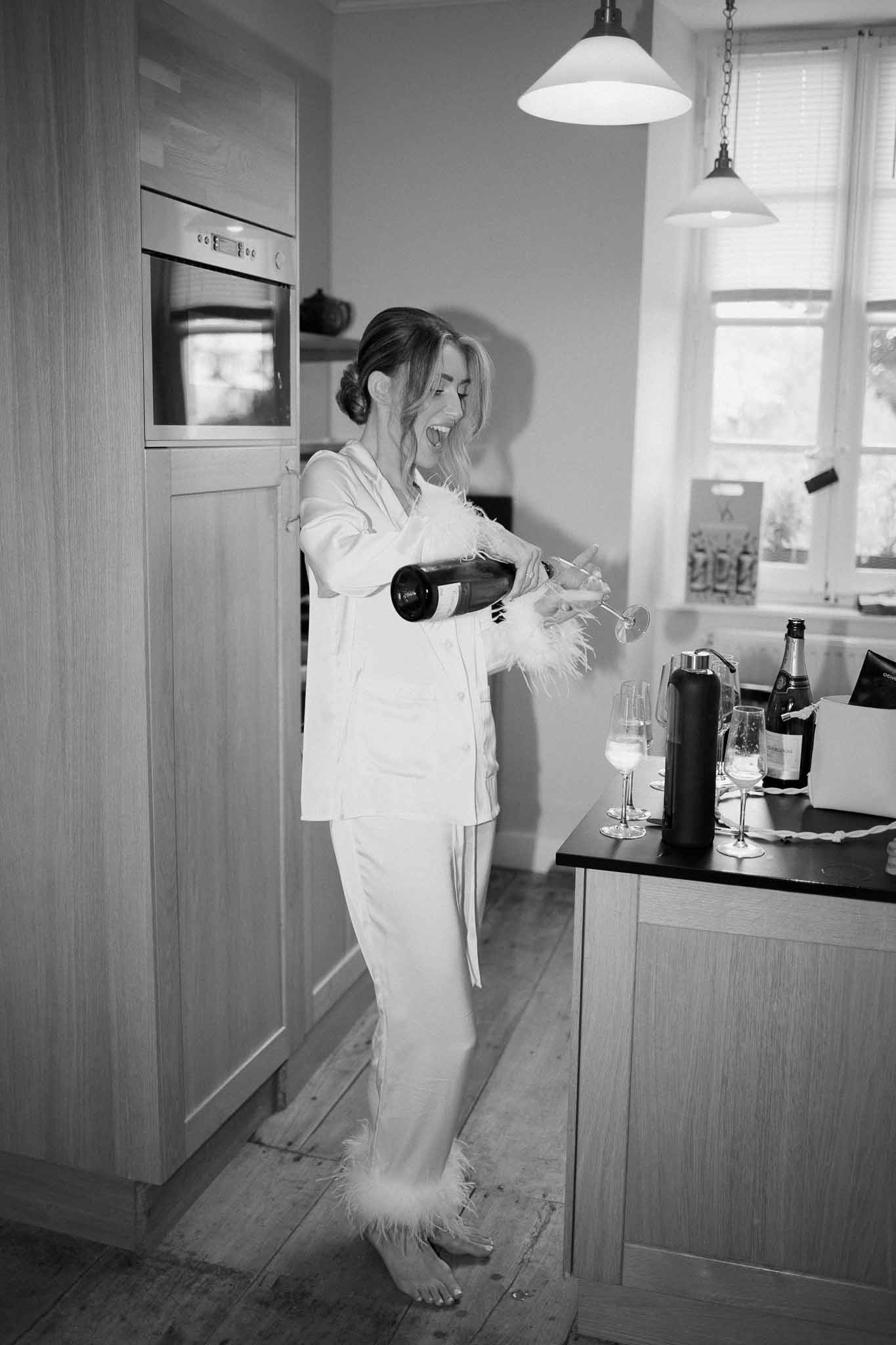 Bride in satin feather-trimmed pajamas pouring champagne in kitchen, laughing, black and white photo