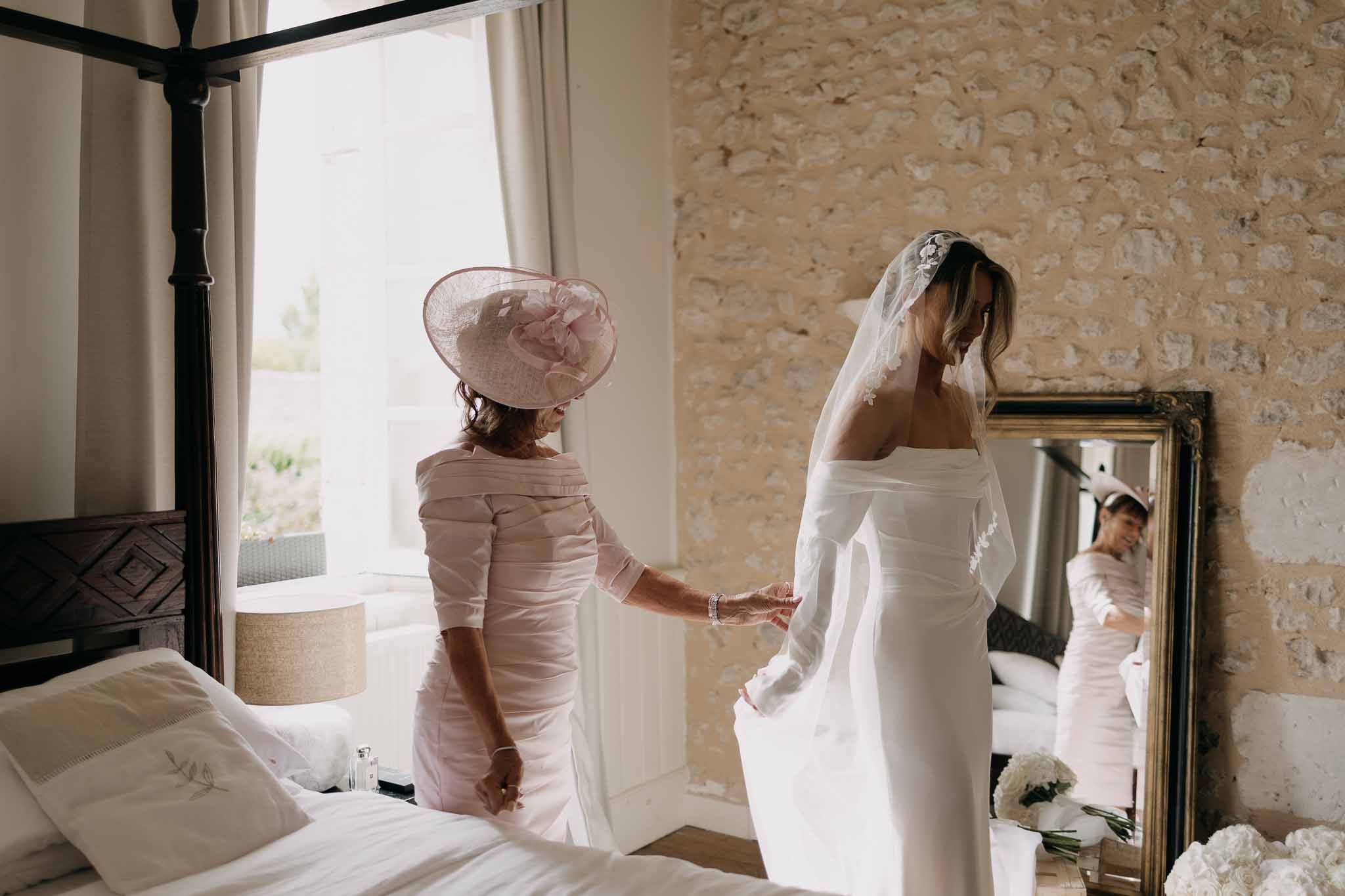 Bride in off-the-shoulder lace-edged veil with mother adjusting dress in chateau bedroom with gilt mirror