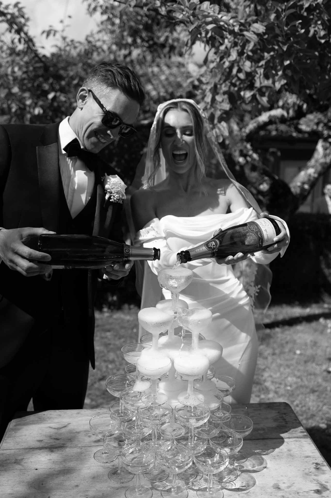 Bride and groom laughing while pouring champagne into a coupe tower in black and white