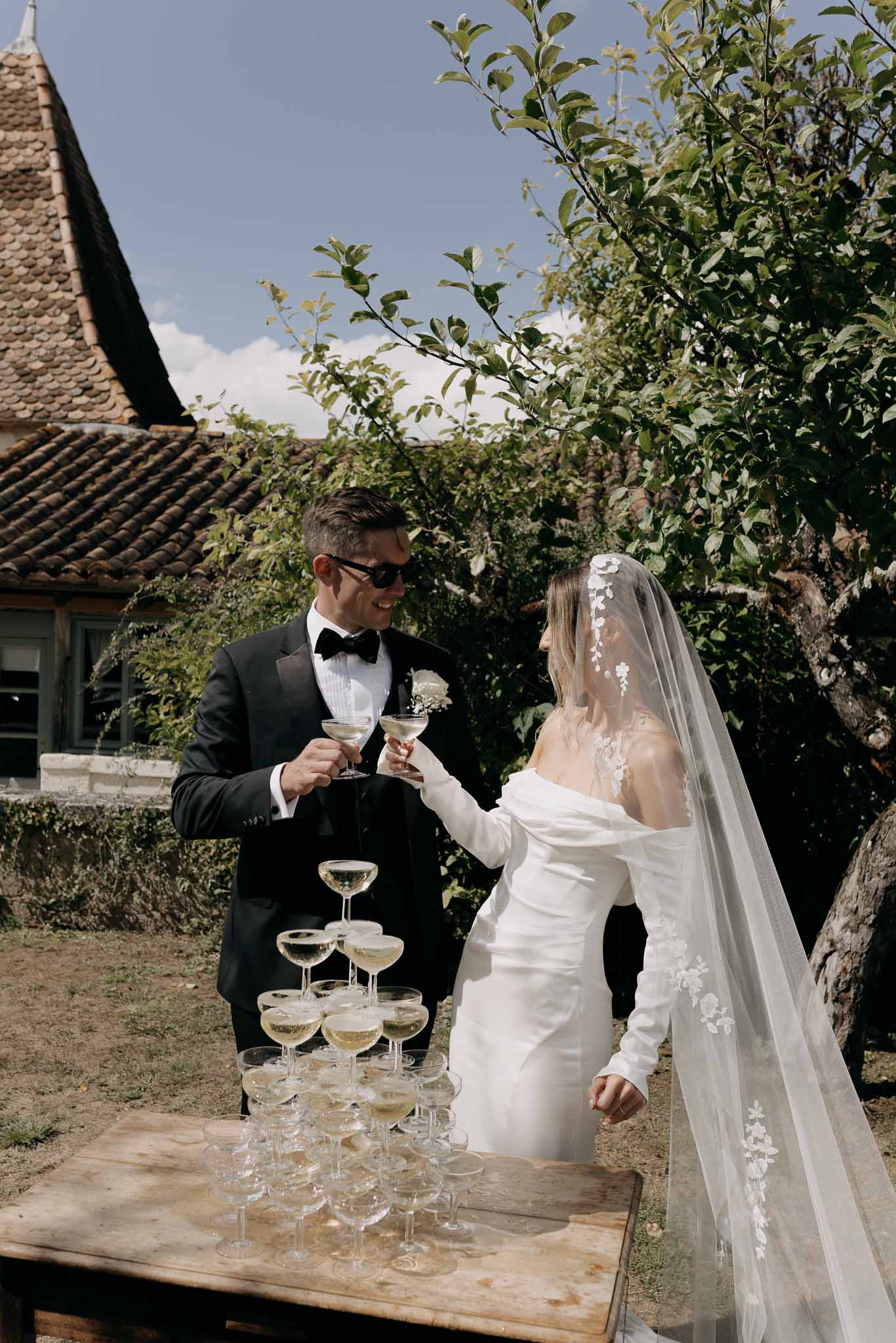 Bride and groom toasting with coupe glasses beside champagne tower at outdoor cocktail hour
