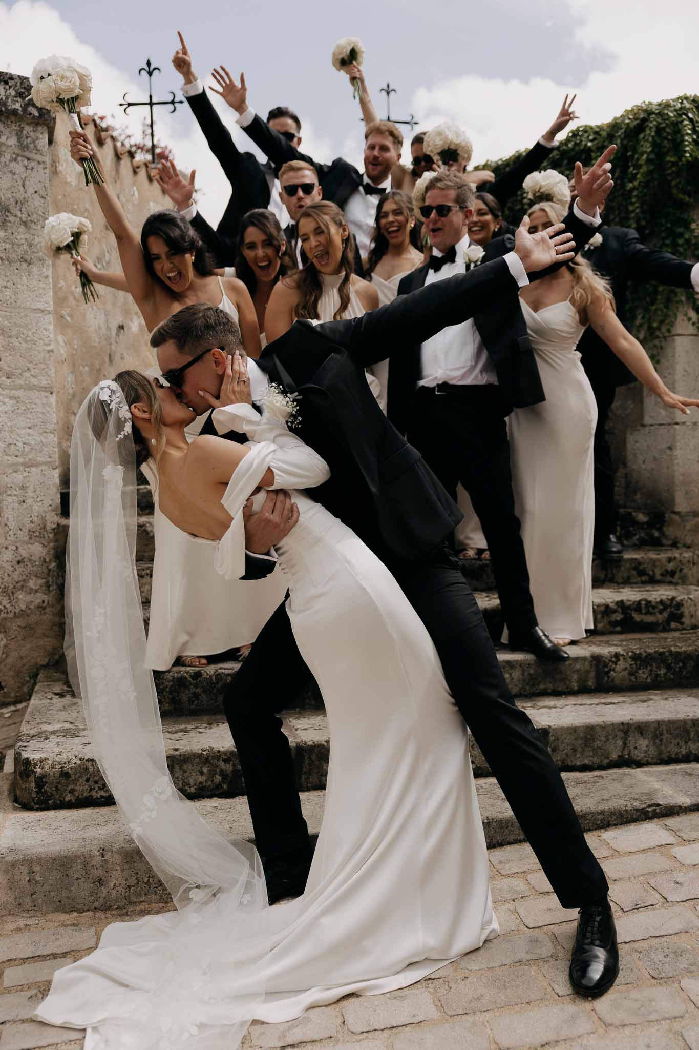 Groom dips and kisses bride on stone steps as bridal party in black tuxedos and ivory gowns cheer behind them