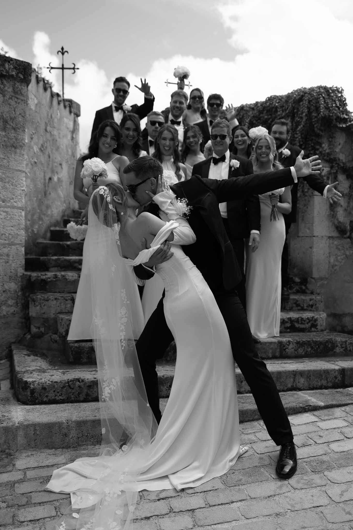 Black and white photo of bride and groom dip kiss on stone steps with bridal party cheering behind them