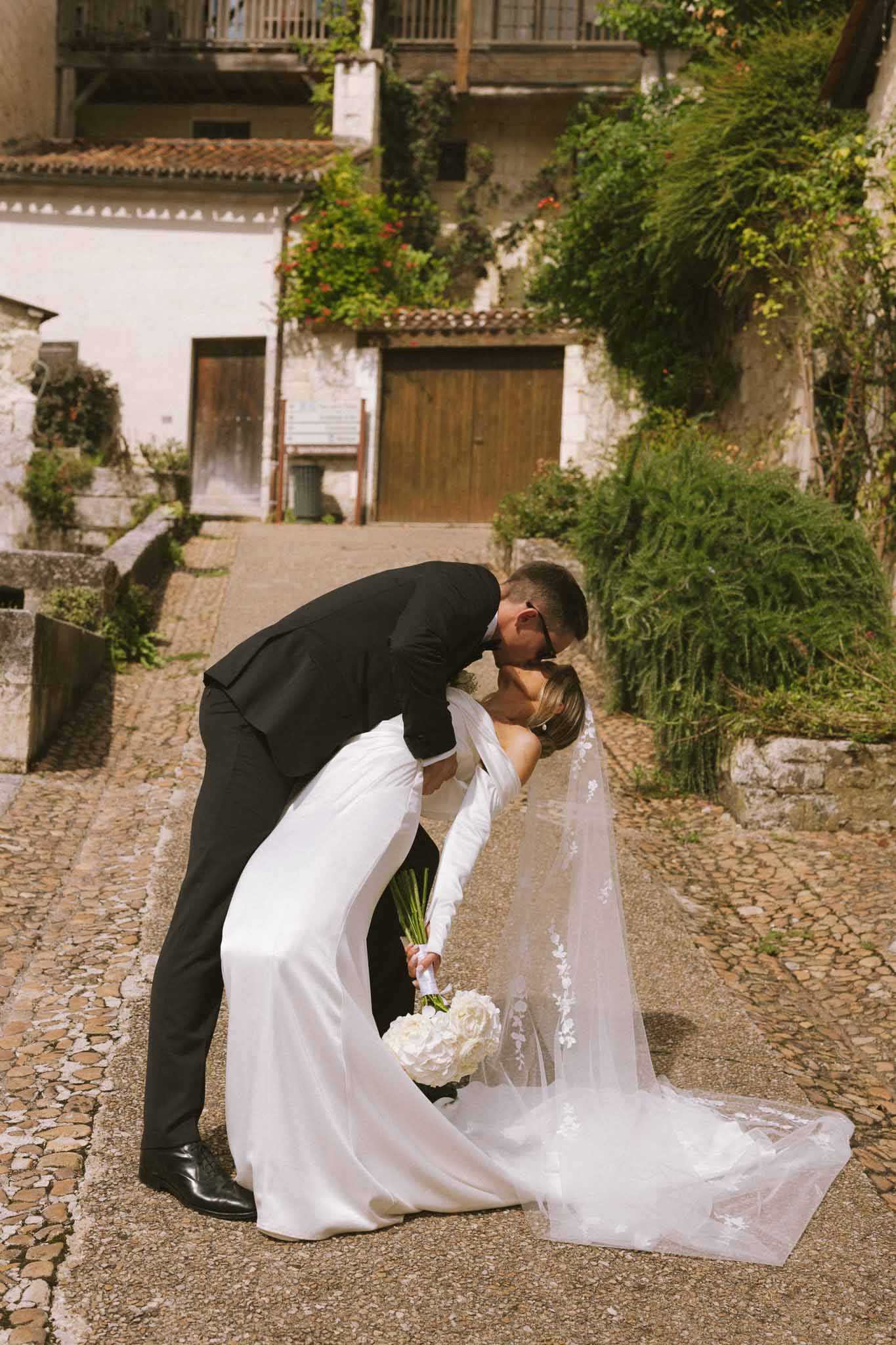 Groom dips bride for a kiss on cobblestone courtyard with cathedral-length veil trailing across stones