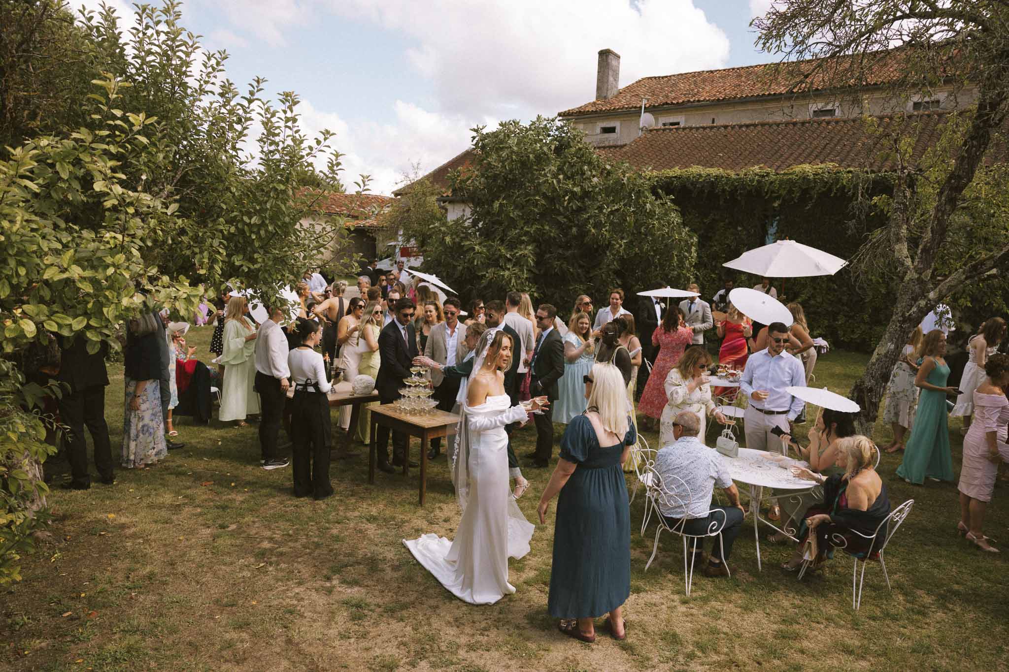 Bride in off-shoulder gown near champagne tower as 55 guests mingle under parasols at ivy-covered manor