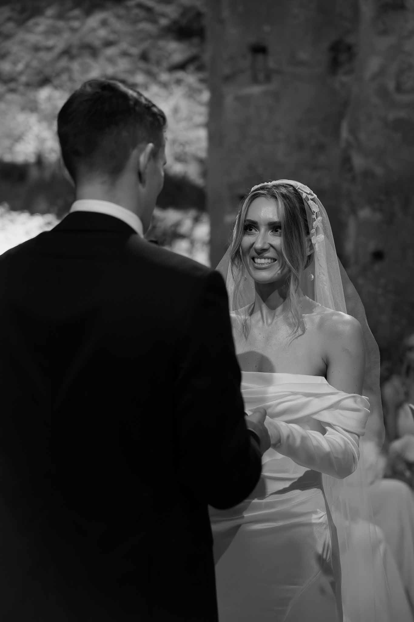 Black and white close-up of bride smiling at groom during ceremony in stone chapel interior