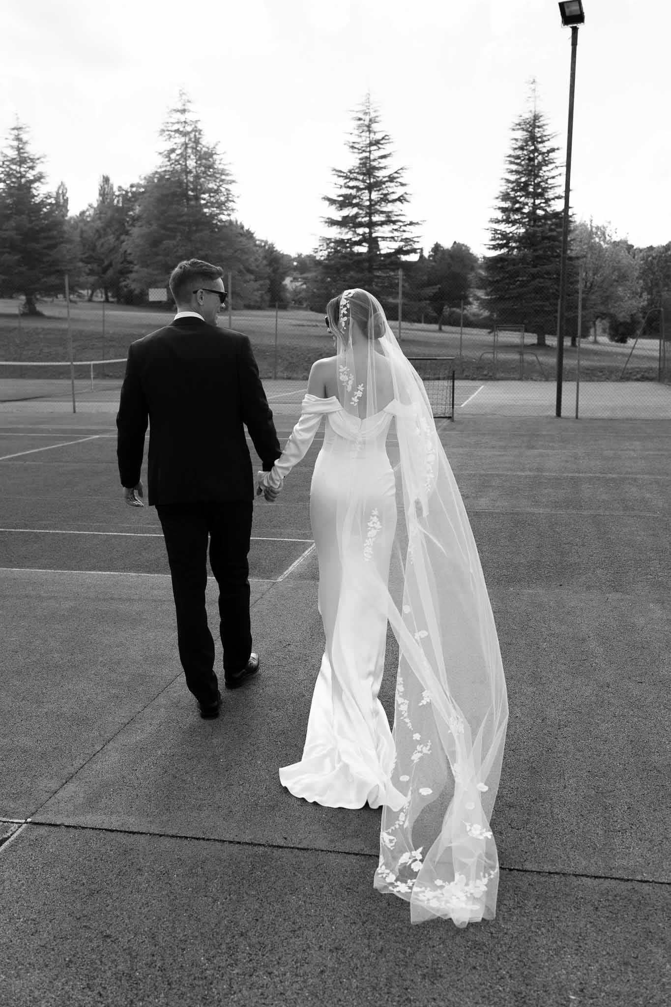 Black and white rear view of couple walking across tennis court with lace-trimmed cathedral veil