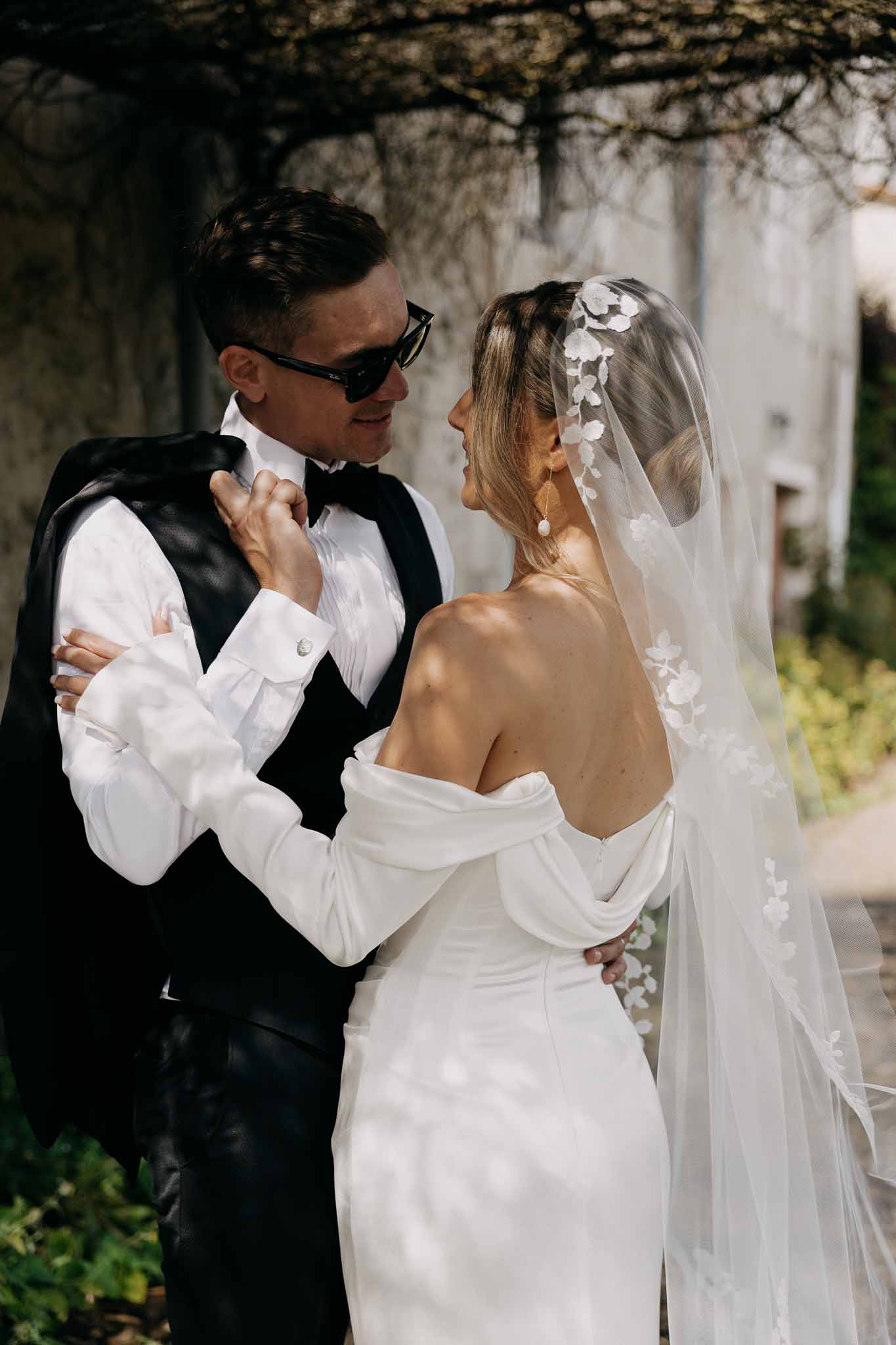 Bride in off-shoulder white gown with lace-edged veil and groom in black tuxedo embracing outdoors