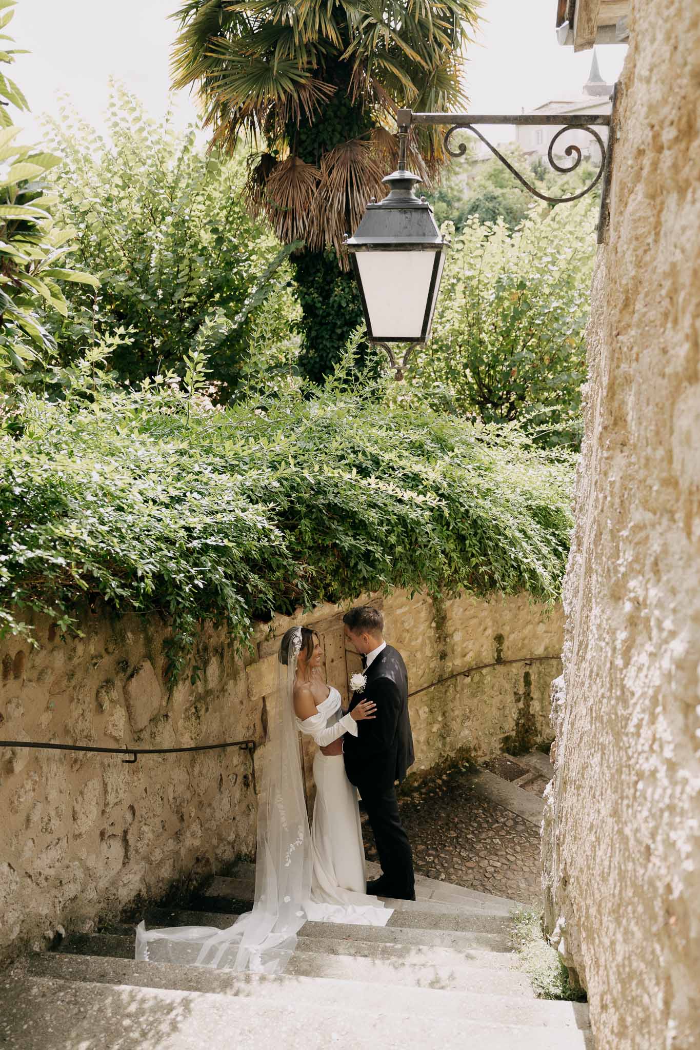 Couple kissing on narrow stone staircase with wrought-iron lantern and climbing greenery in French village alley