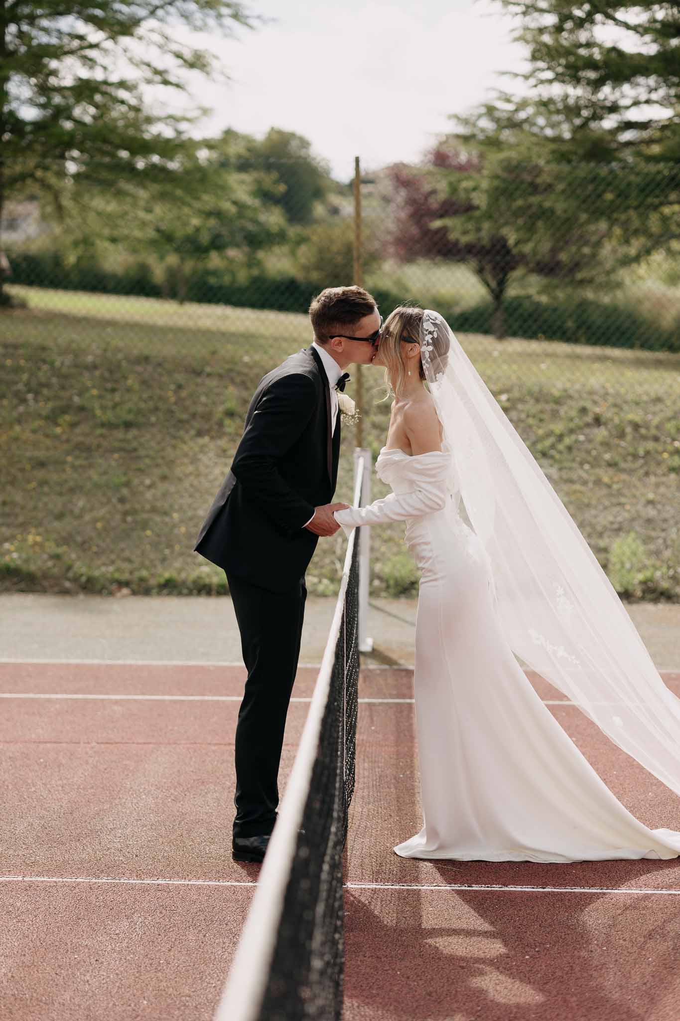 Bride and groom in sunglasses kissing across a tennis court net in a playful fashion-forward portrait