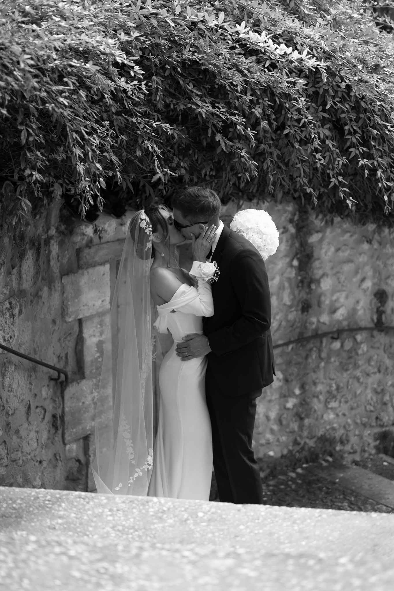 Black and white couple kissing before stone arch with climbing foliage bride in off-shoulder gown with floral-trimmed veil