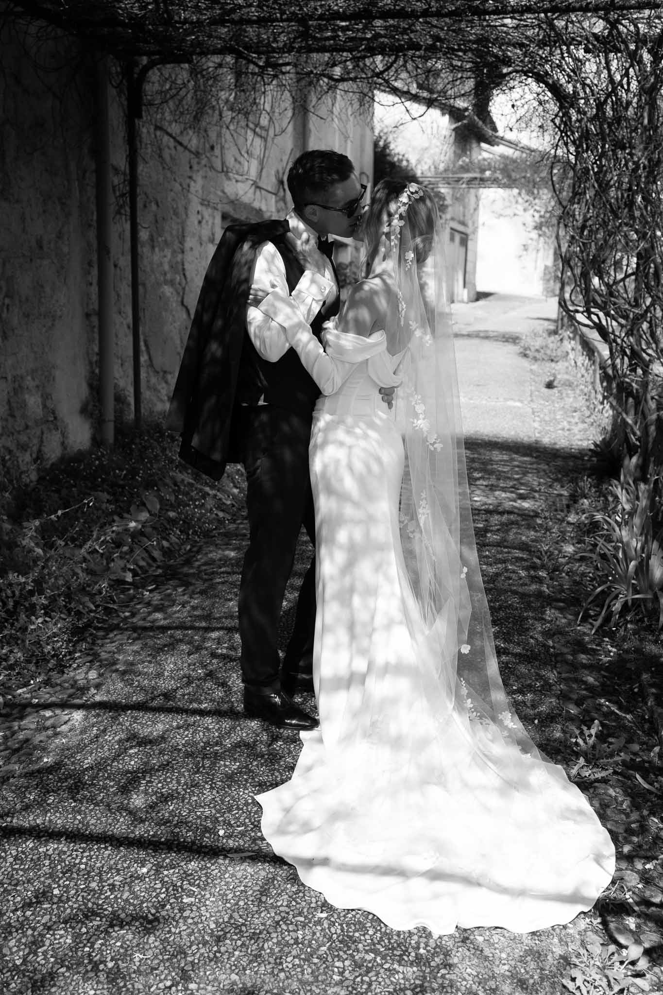 Black and white portrait of bride and groom kissing under vine-covered pergola with cathedral veil trailing on gravel path