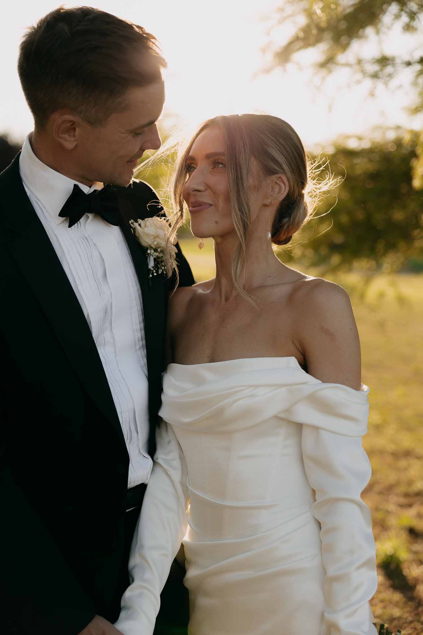 Close-up couple portrait at golden hour, groom in tuxedo and bride in off-shoulder ivory gown with updo