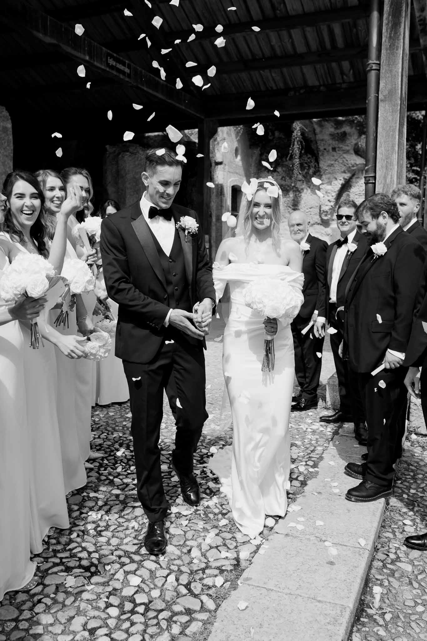 Couple exits stone chapel through flower petal toss lined by bridesmaids and groomsmen on cobblestone in B&W
