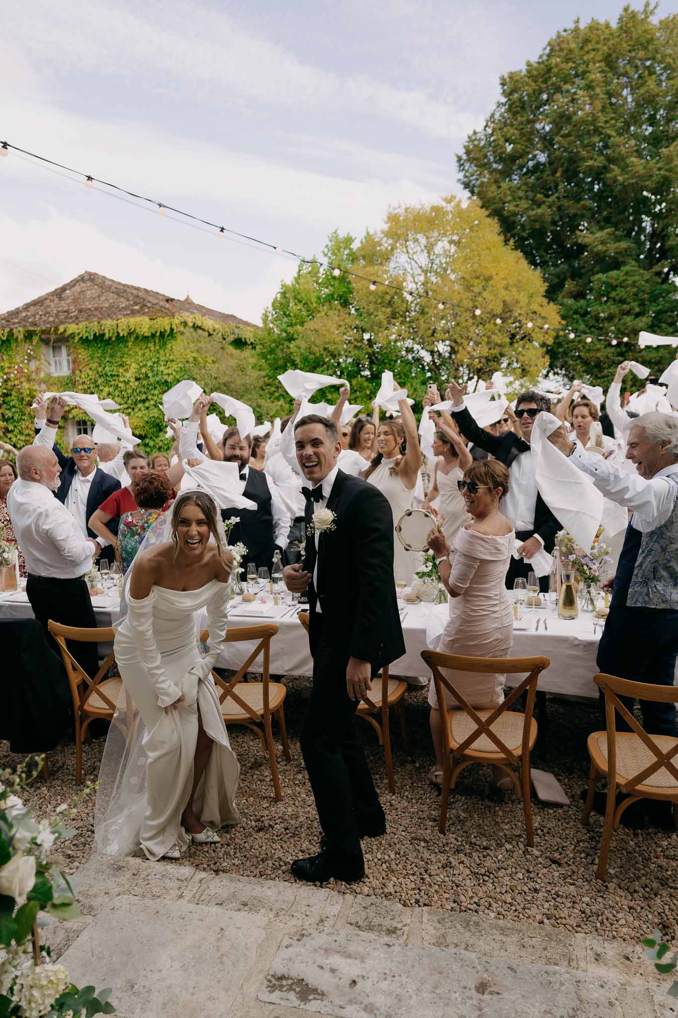 Bride and groom walk past banquet table as guests wave white napkins in celebration at a courtyard reception