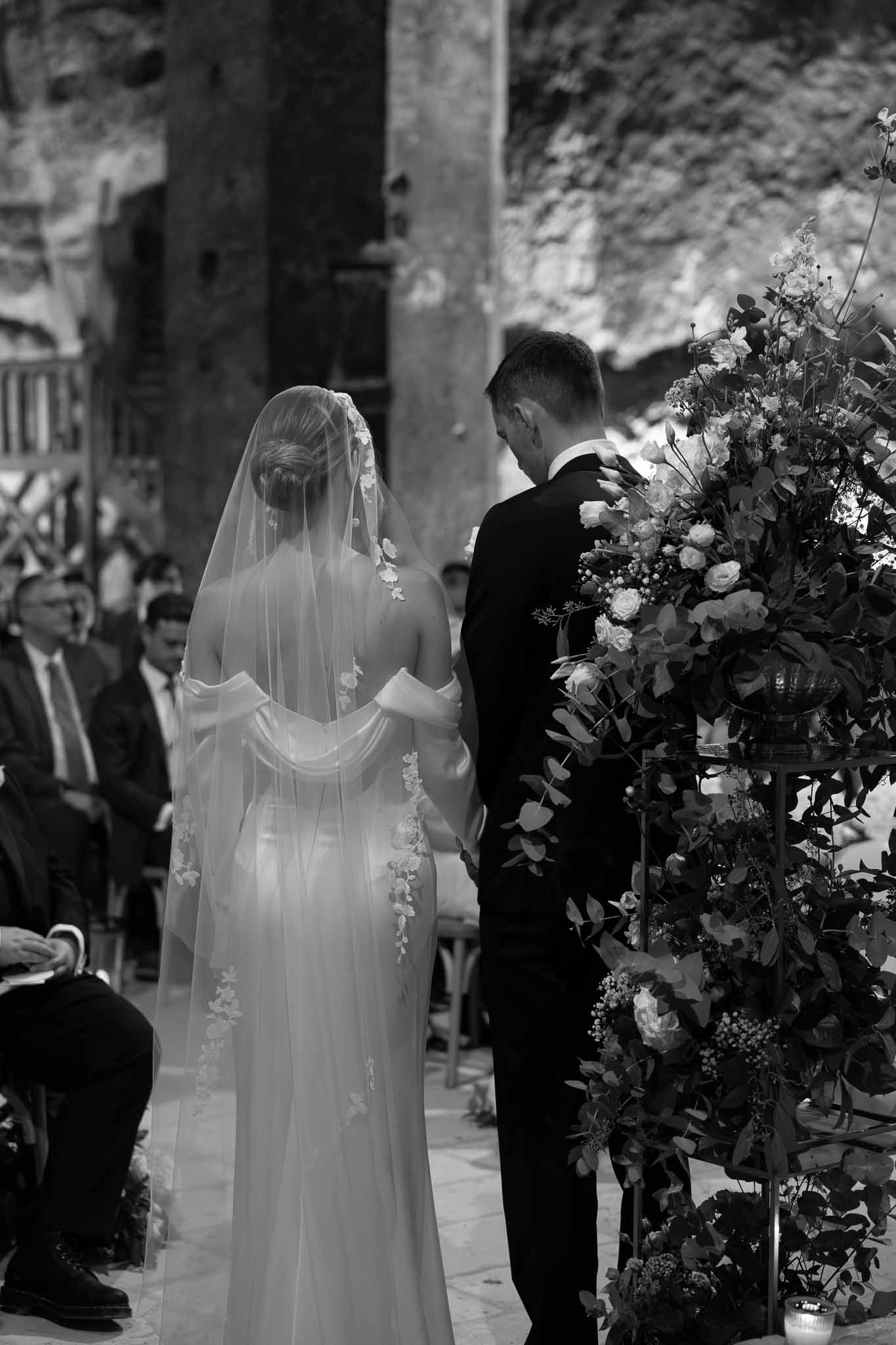Black and white rear view of couple at altar in stone ruin with floral urn and lace-motif veil