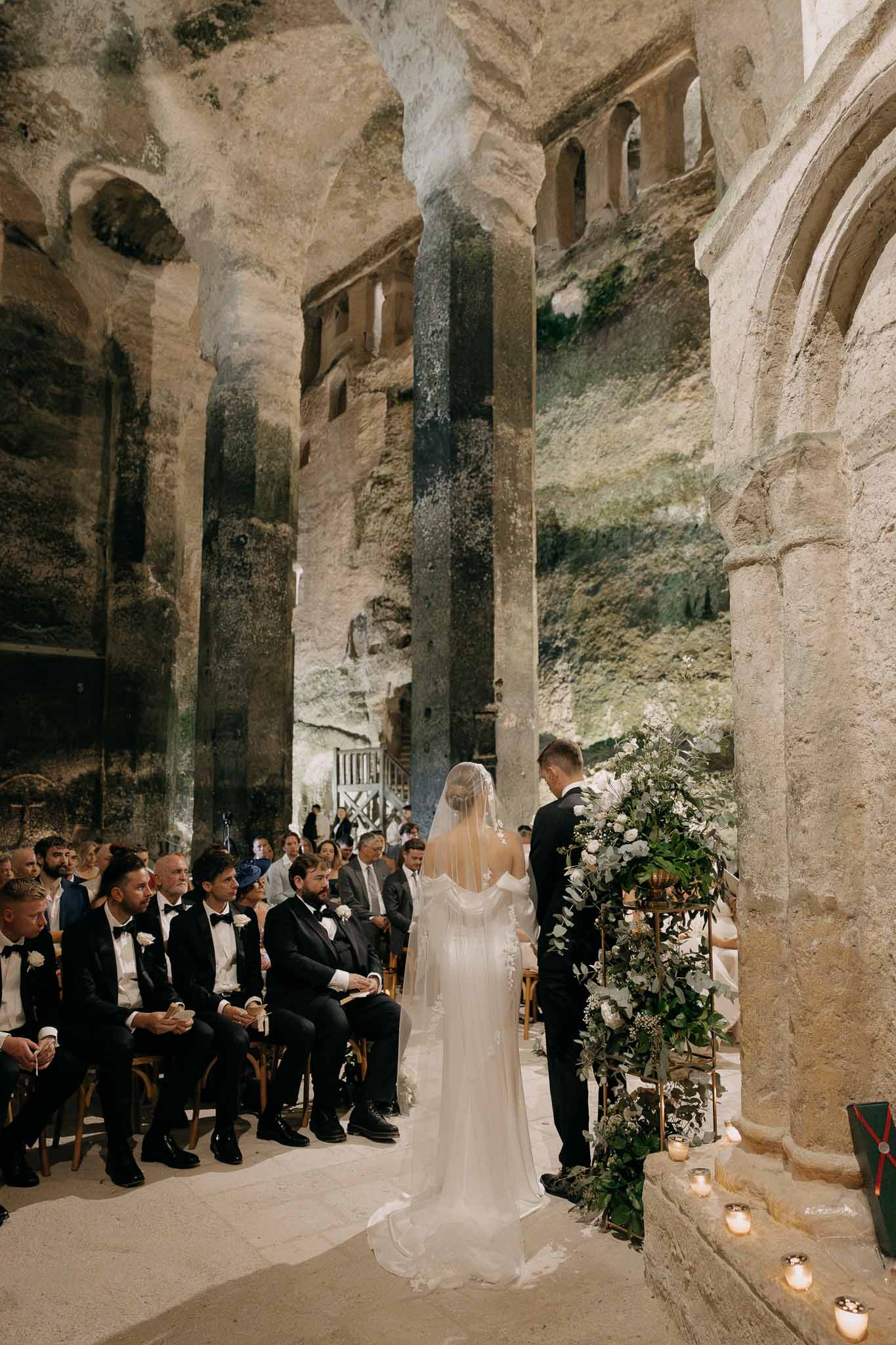 Couple at altar in troglodyte stone chapel with carved columns and white rose arrangement beside candles