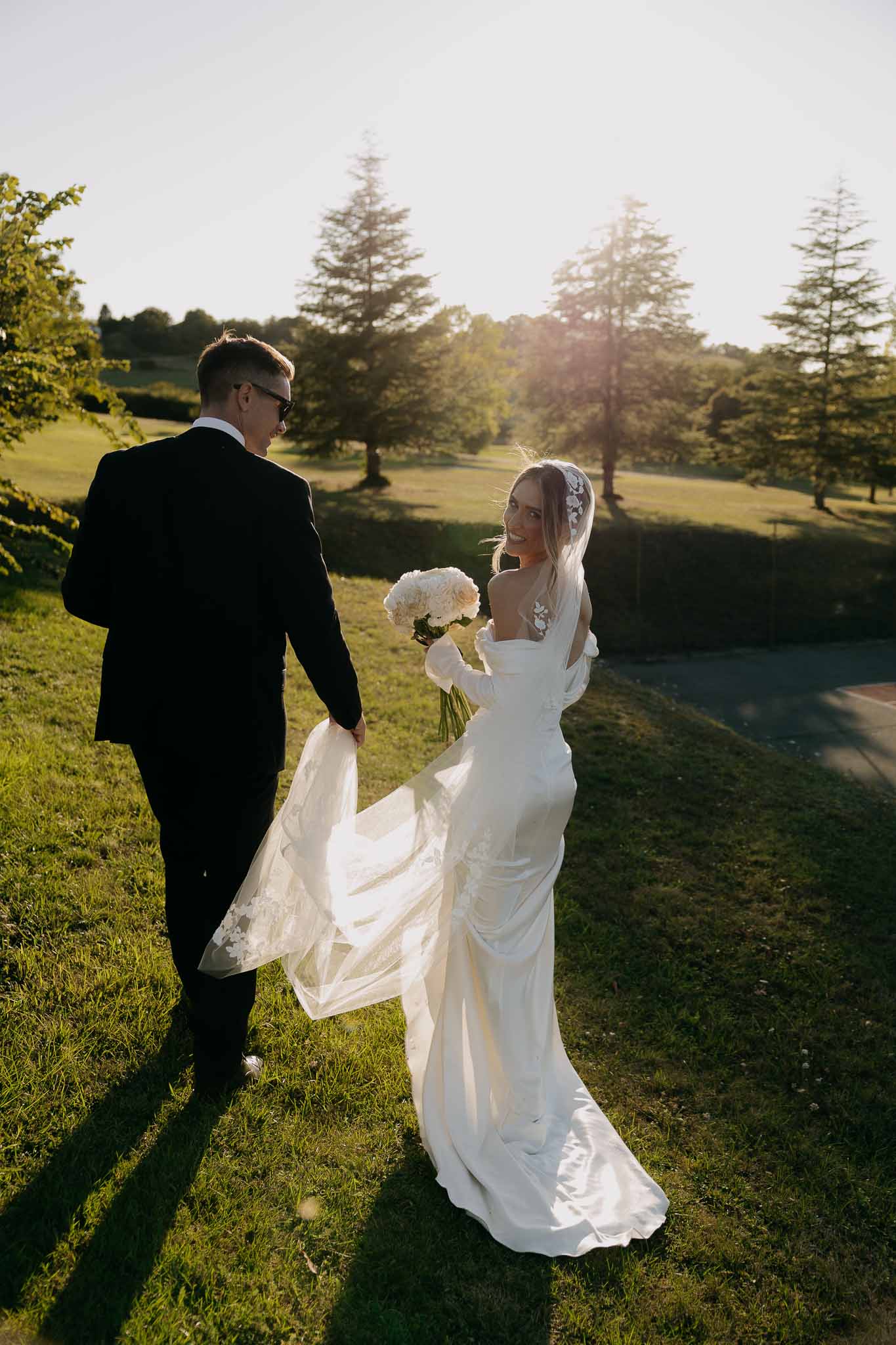 Groom holds bride's lace-trimmed train as they walk on grassy hillside with golden sun flare behind