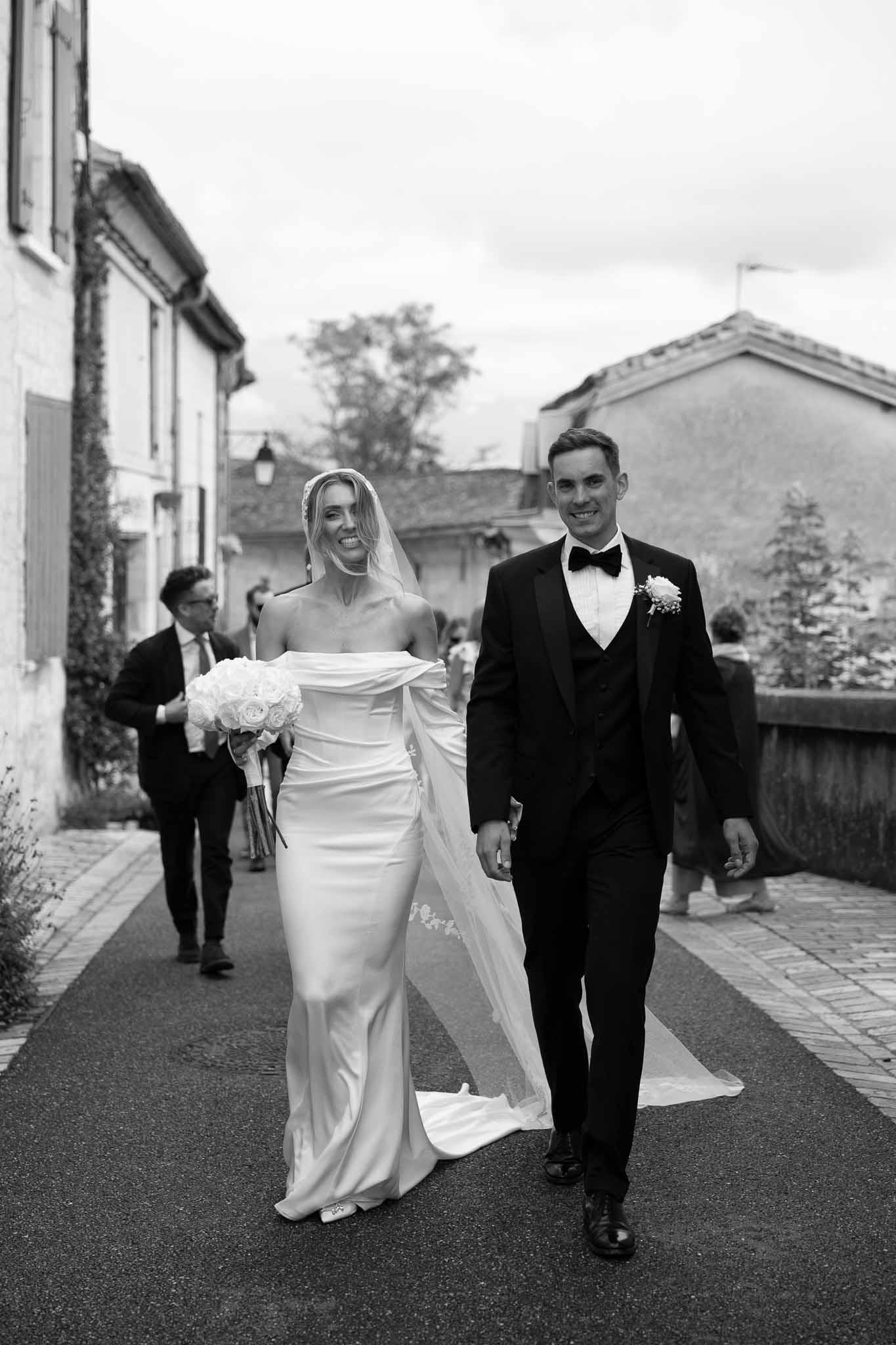 Bride in off-shoulder satin gown with lace-trimmed veil and groom in tuxedo walking village street in B&W