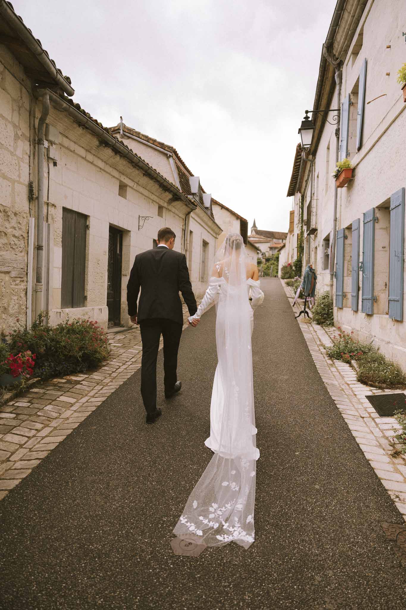 Bride and groom walking hand in hand down a French village street, cathedral-length veil trailing behind them