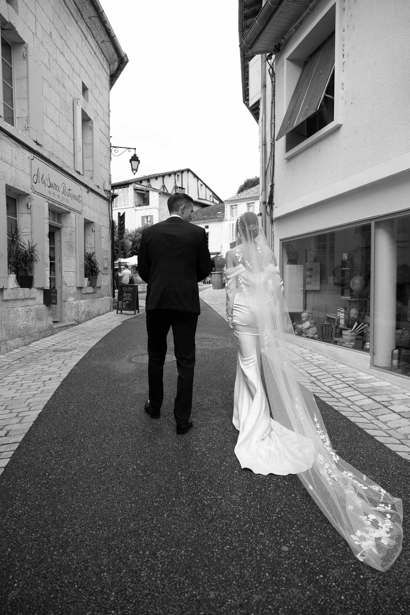 Black and white photo of bride and groom walking down French village street with cathedral-length lace veil trailing behind