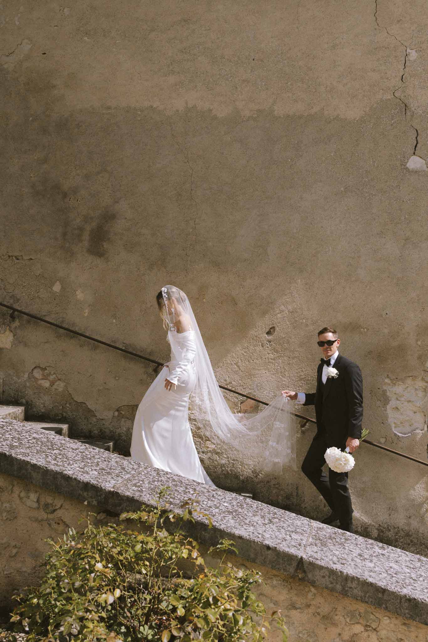 Couple ascending stone staircase as groom holds trailing cathedral veil and white peony bouquet