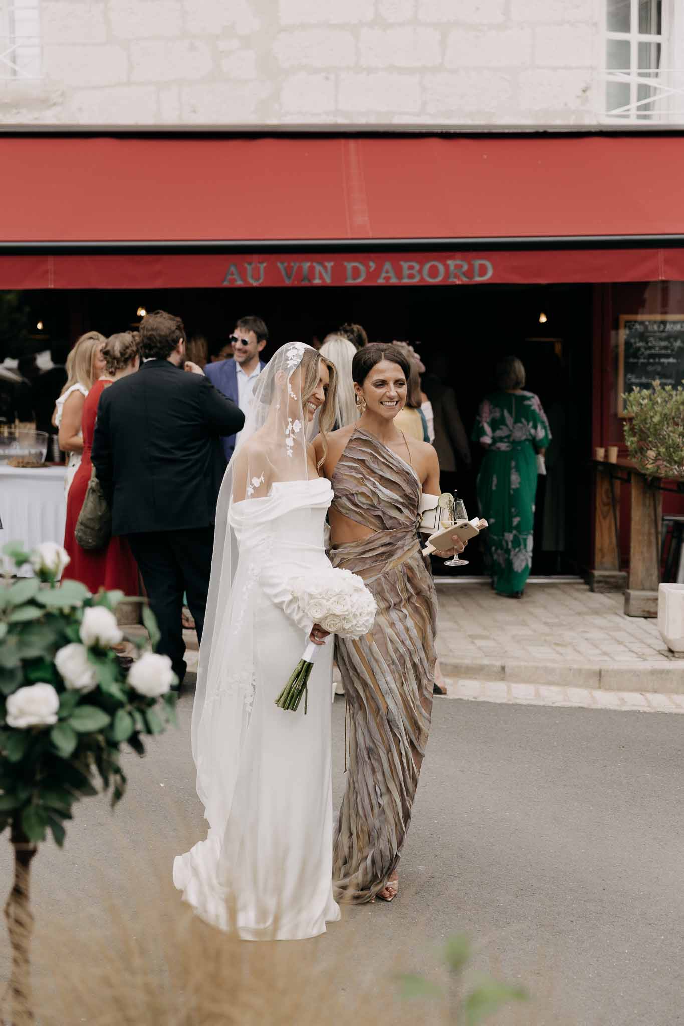 Bride laughing in off-shoulder gown and floral lace veil with white rose bouquet outside wine bar with guests