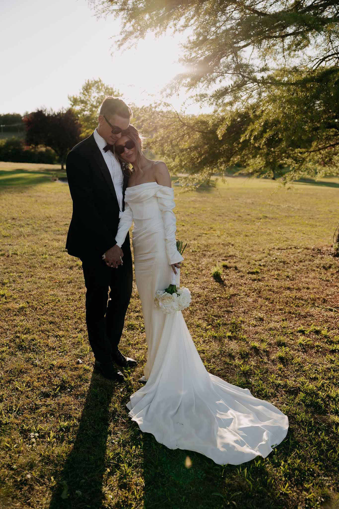 Bride in off-shoulder white gown with sunglasses and groom in black tuxedo backlit at golden hour in parkland