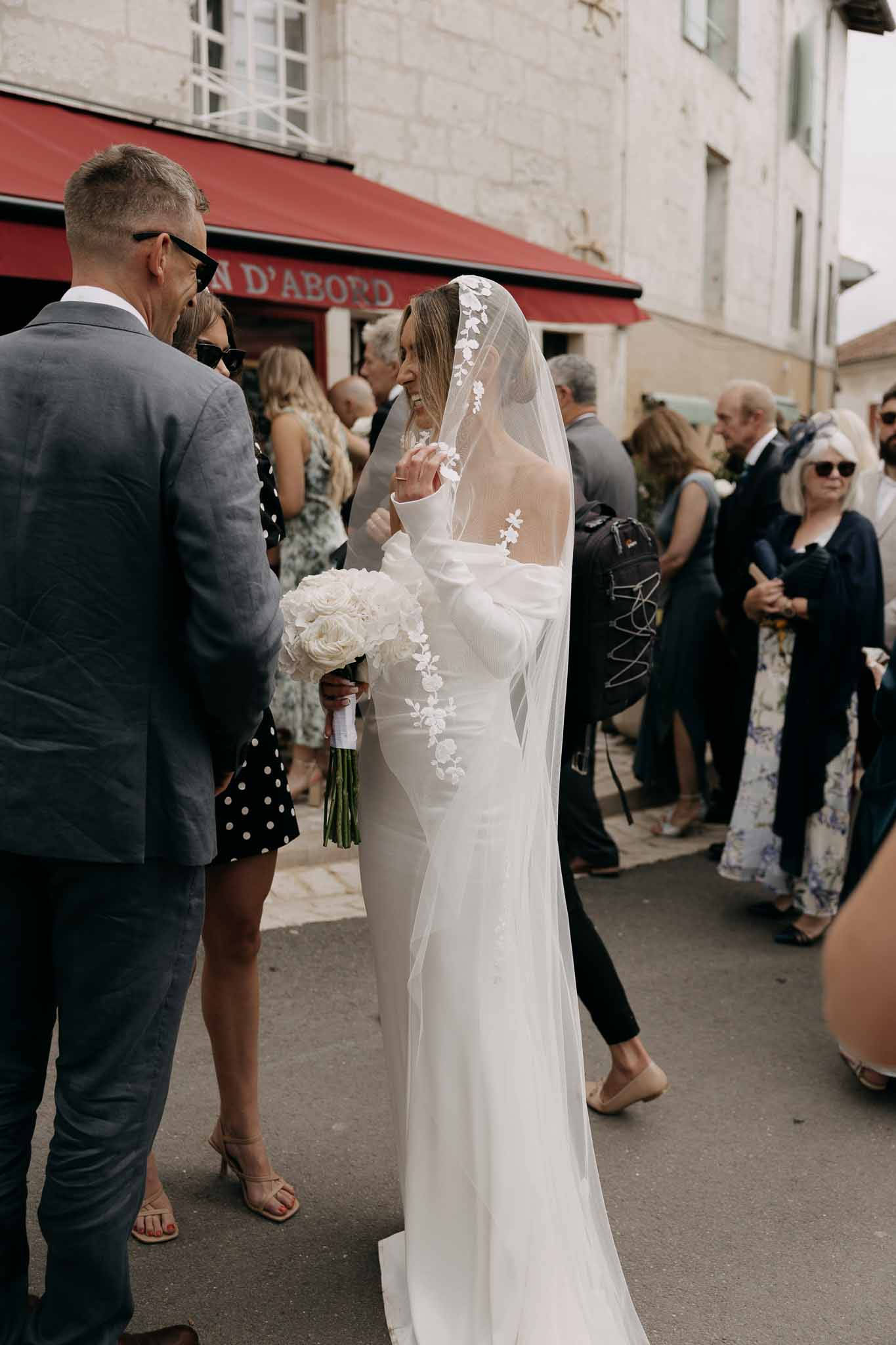 Bride in fitted ivory gown with lace cathedral veil holding white ranunculus bouquet greeting guests on street