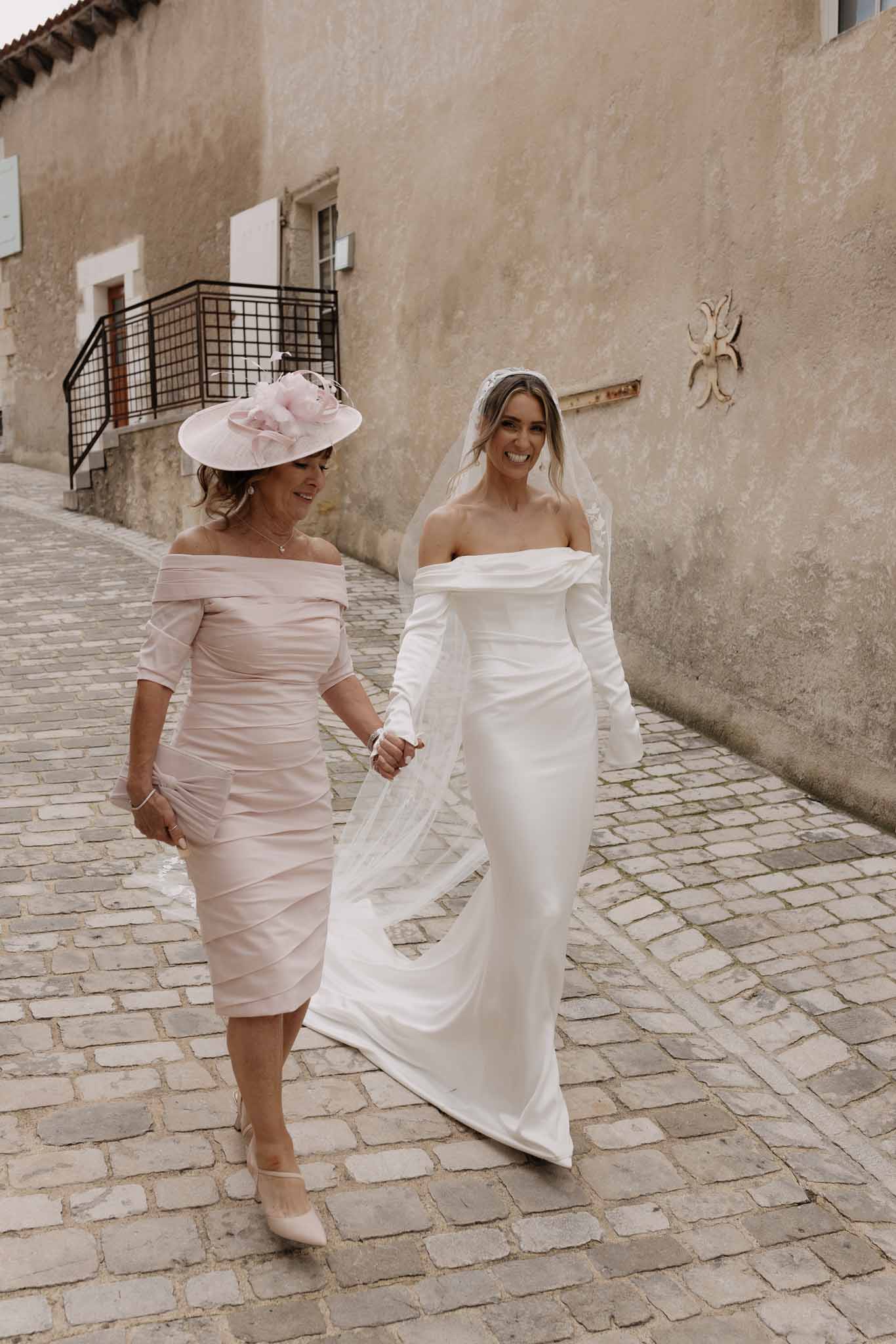 Bride and mother walking hand in hand along cobblestone alley in white gown and blush dress