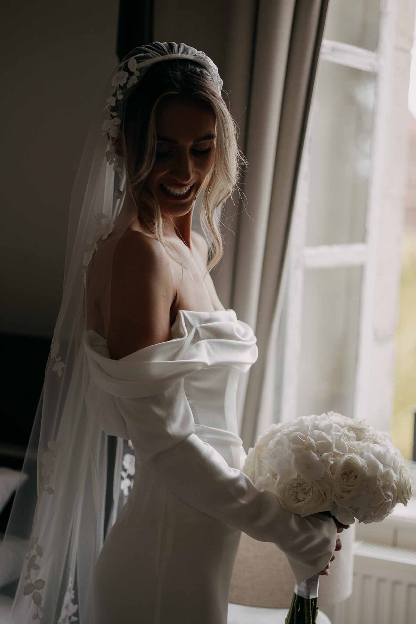 Bride laughing by window in ivory off-shoulder satin gown with floral veil headband and white peony bouquet