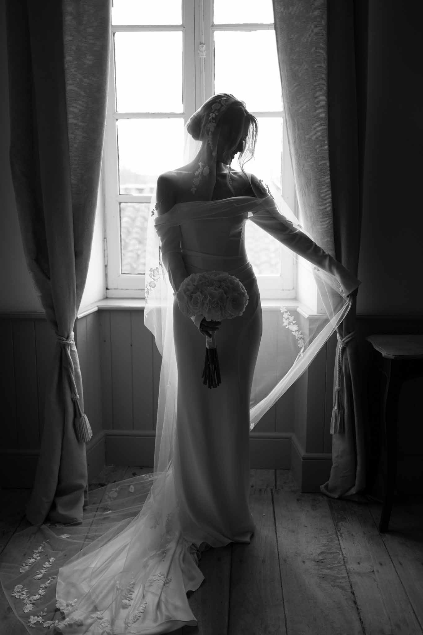Black-and-white bridal portrait from behind at tall window, cathedral train and lace-edged veil extended