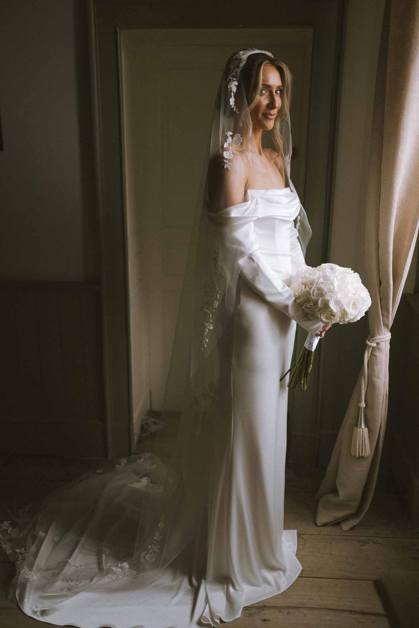 Bride in off-the-shoulder satin gown with lace-trimmed veil holding ivory rose bouquet in a moody chateau room