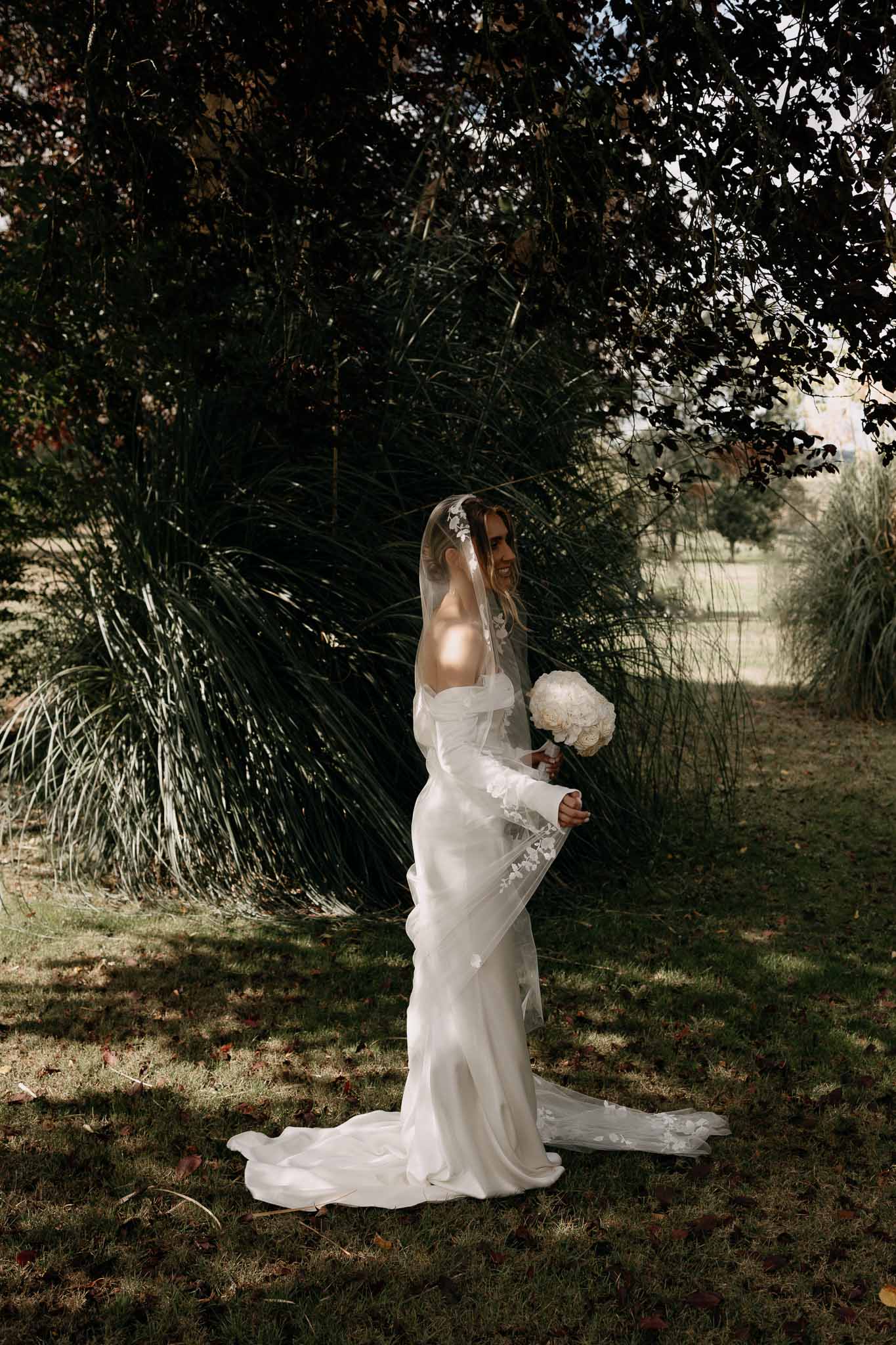 Bride in satin gown with lace bell-sleeve overlay and applique veil holding white peony bouquet